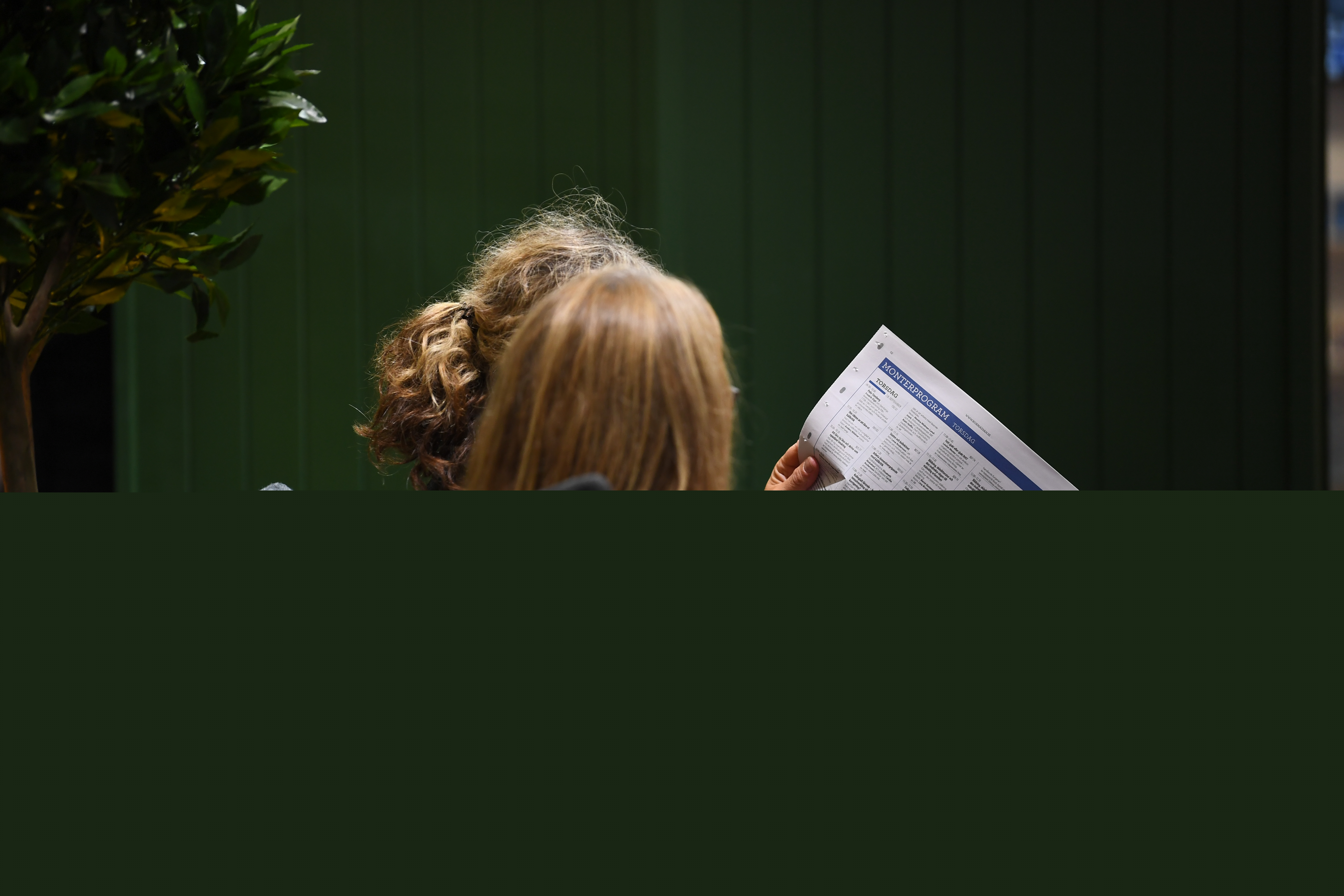 Två kvinnor sitter bredvid varandra inomhus och läser en tidning.