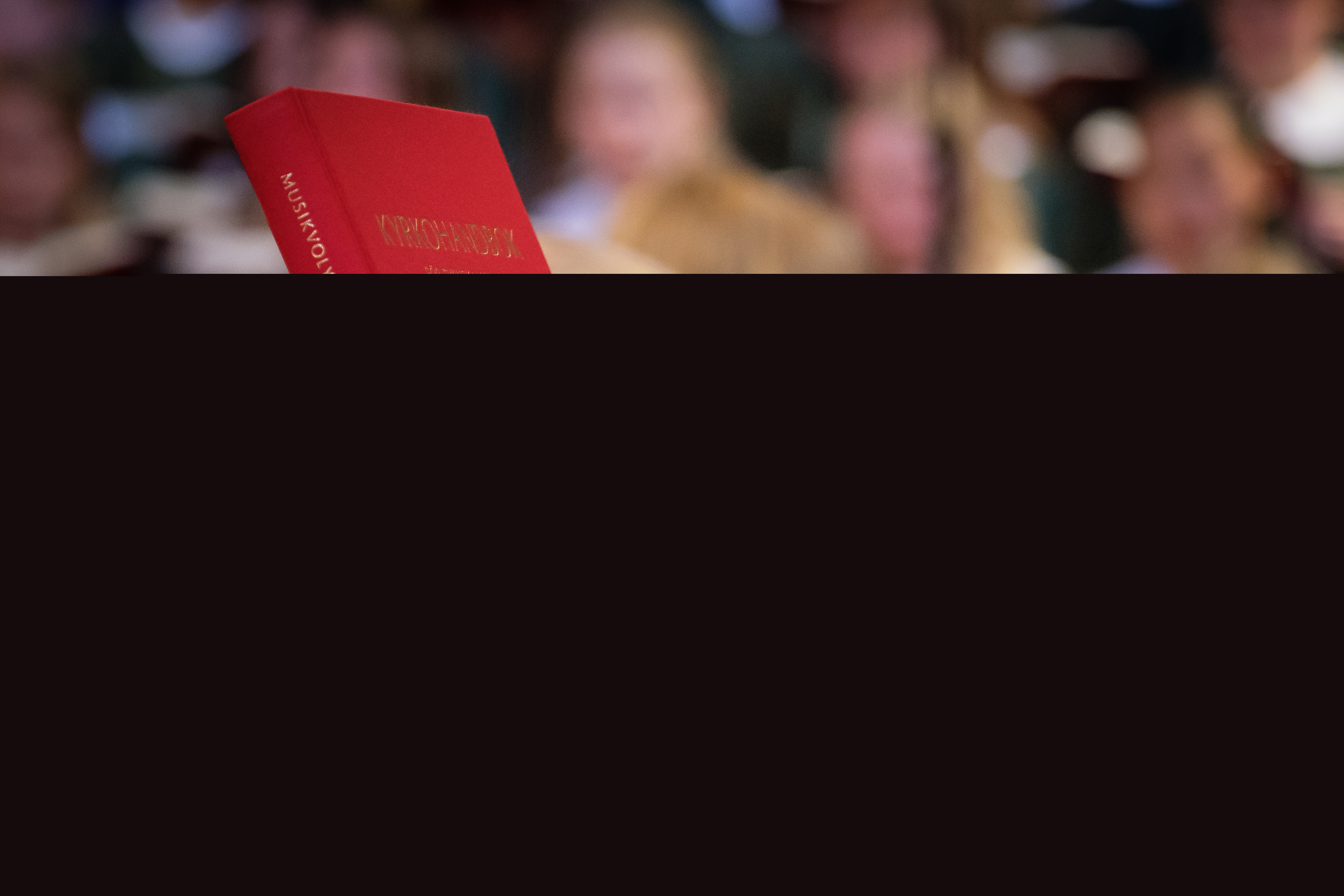 Två personer håller upp kyrkohandboken i luften. En flickkör syns suddigt i bakgrunden.