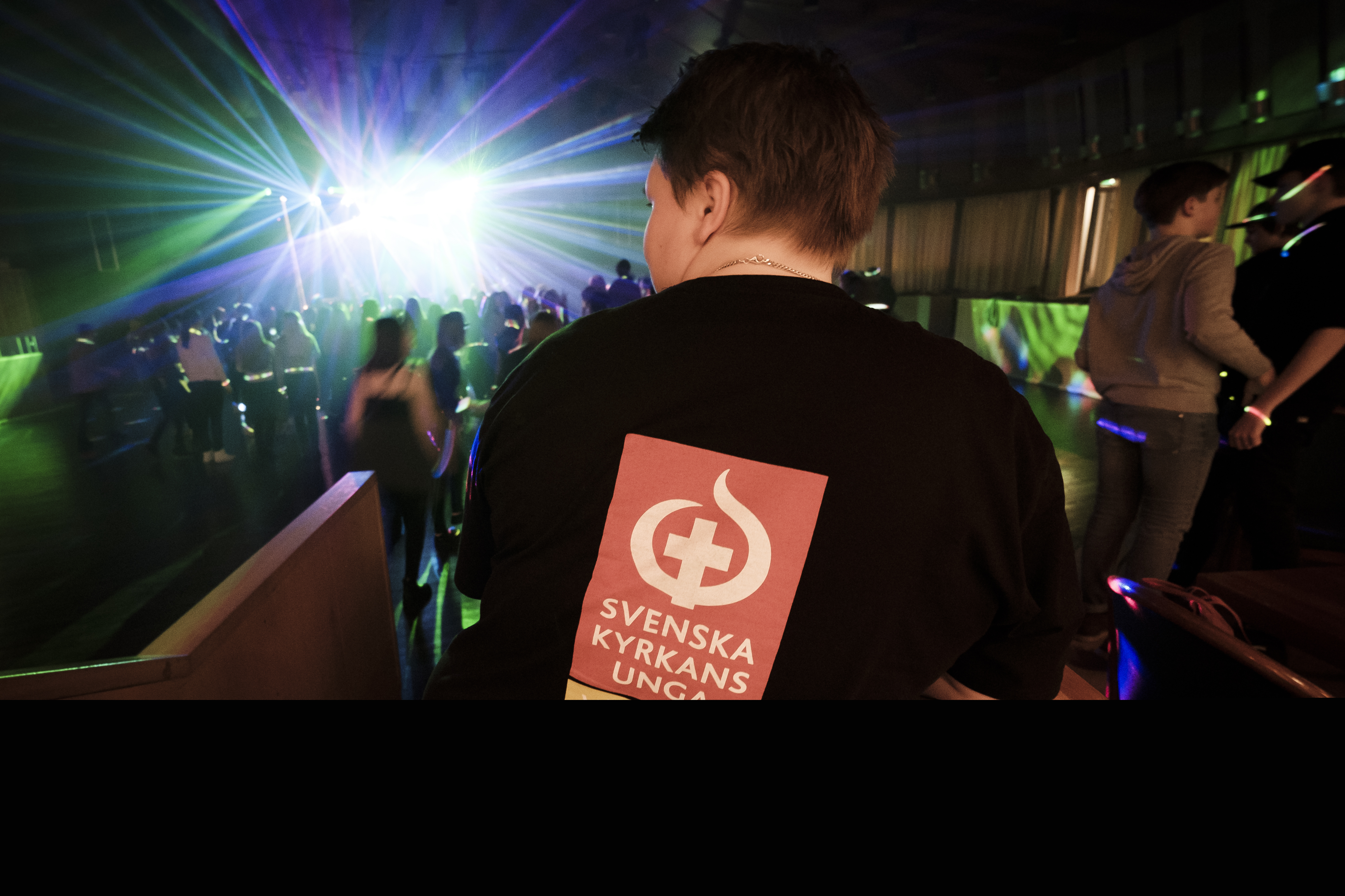 En ungdom har en tröja med texten Svenska kyrkans unga på ryggen. I bakgrunden syns ett fullt dansgolv med discoljus.