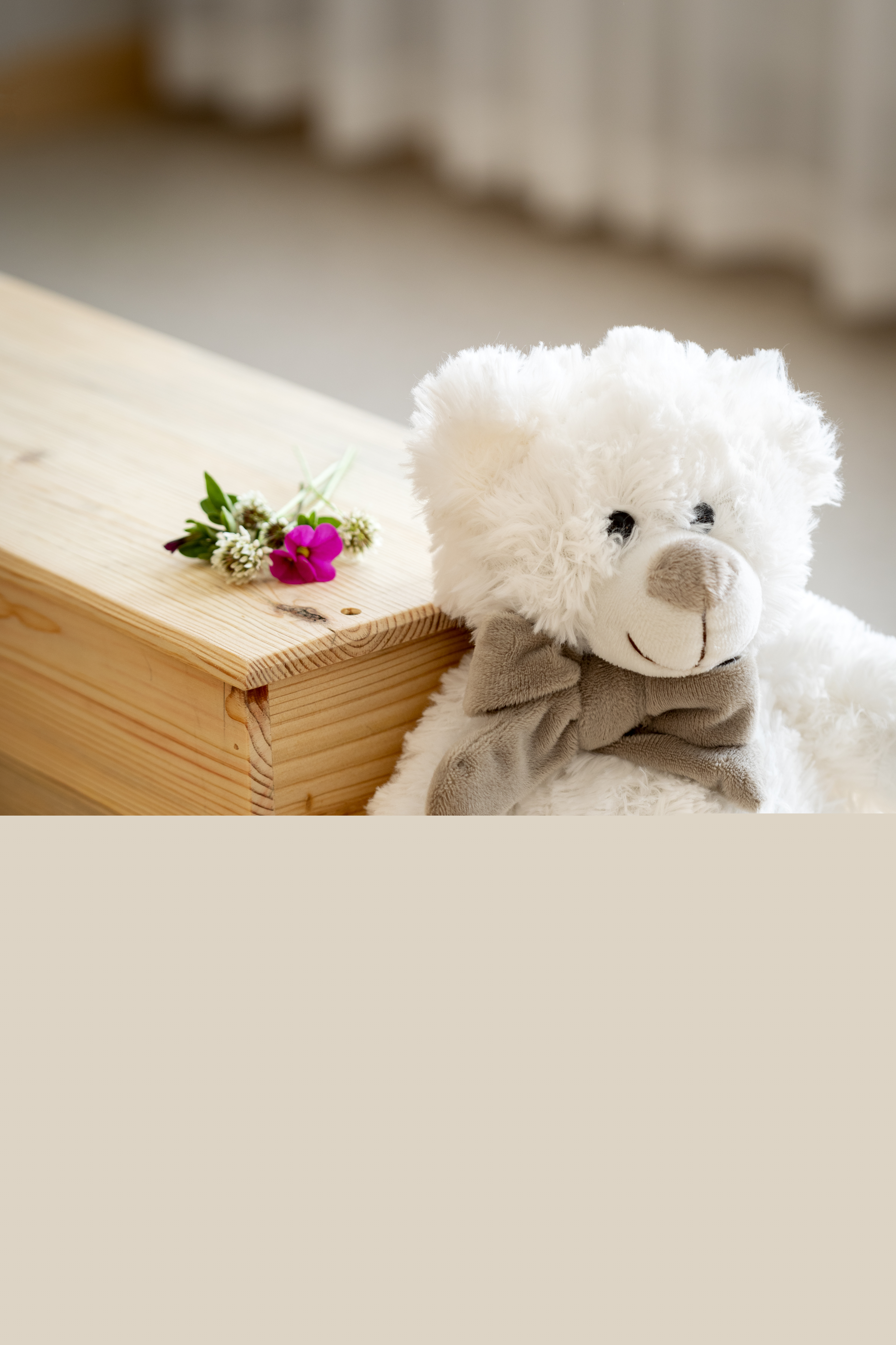 En vit nallebjörn sitter lutad mot en liten träkista. Några blommor ligger på kistan.