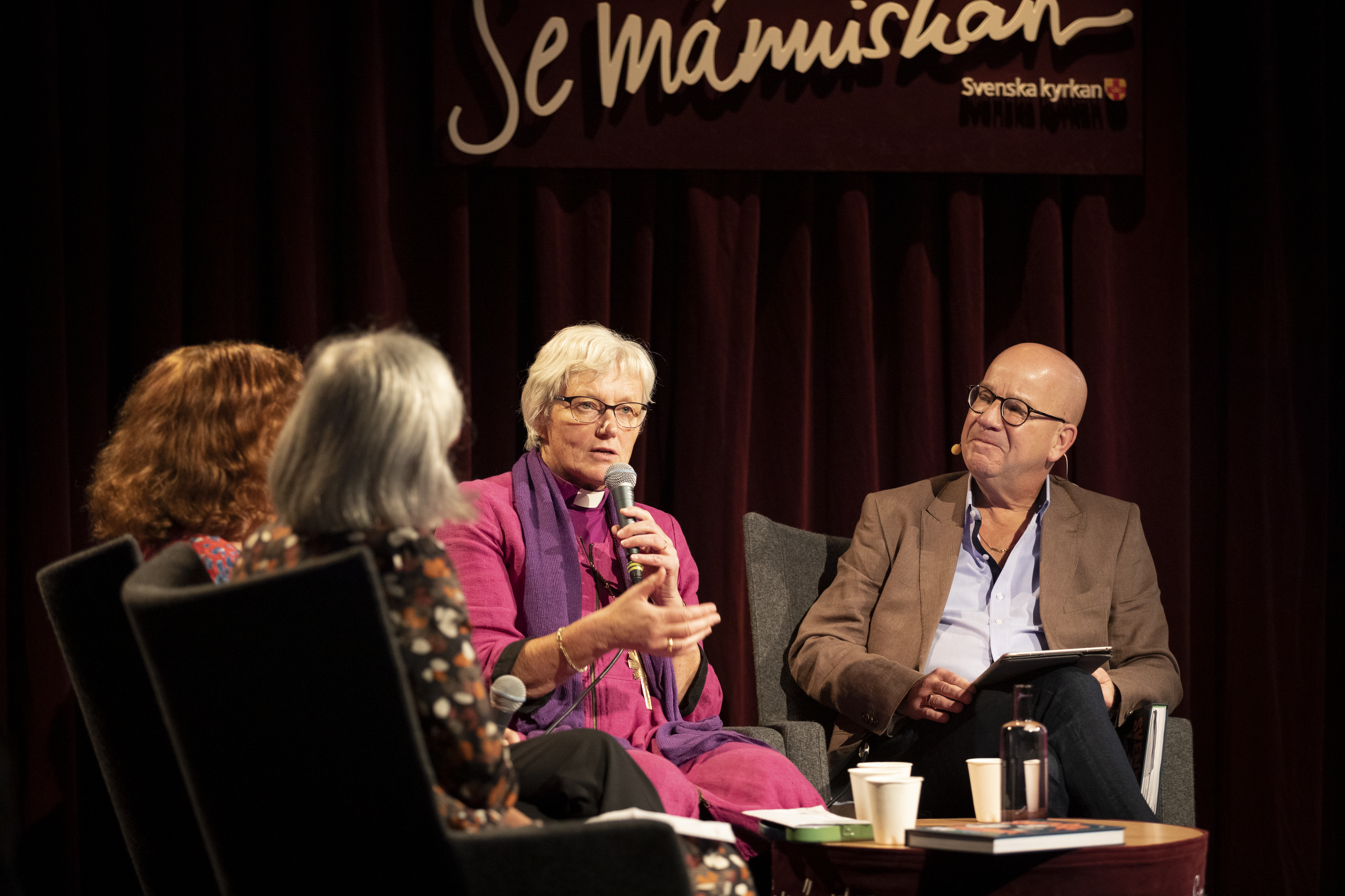 Ärkebiskop Antje Jackelén sitter i en studiomiljö och pratar i en mikrofon. På väggen bakom står det Se människan.
