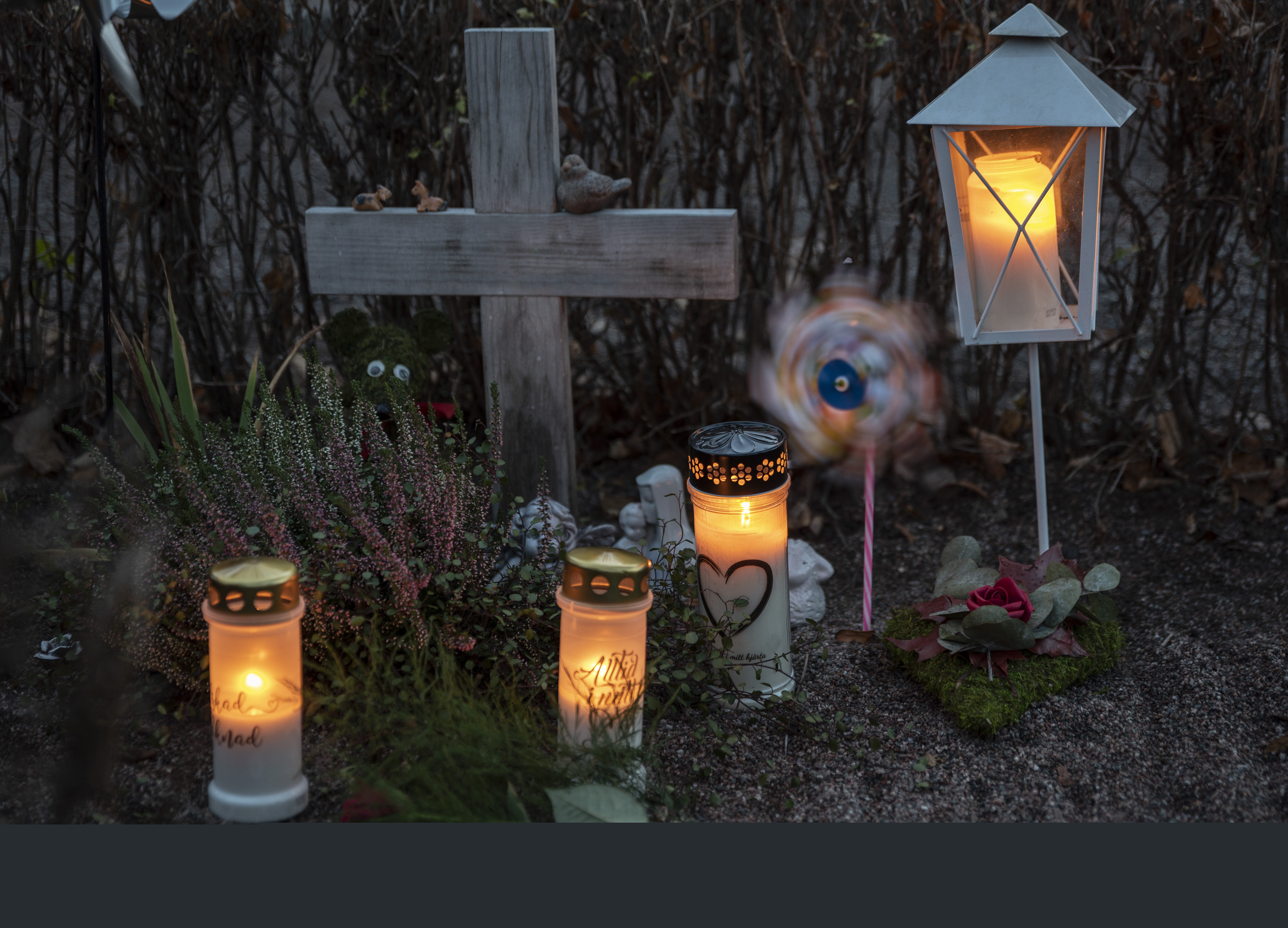 En grav med ett enkelt träkors har smyckats med gravljus, en lykta, en vindsnurra, ler- och gipsfigurer samt olika blommor.