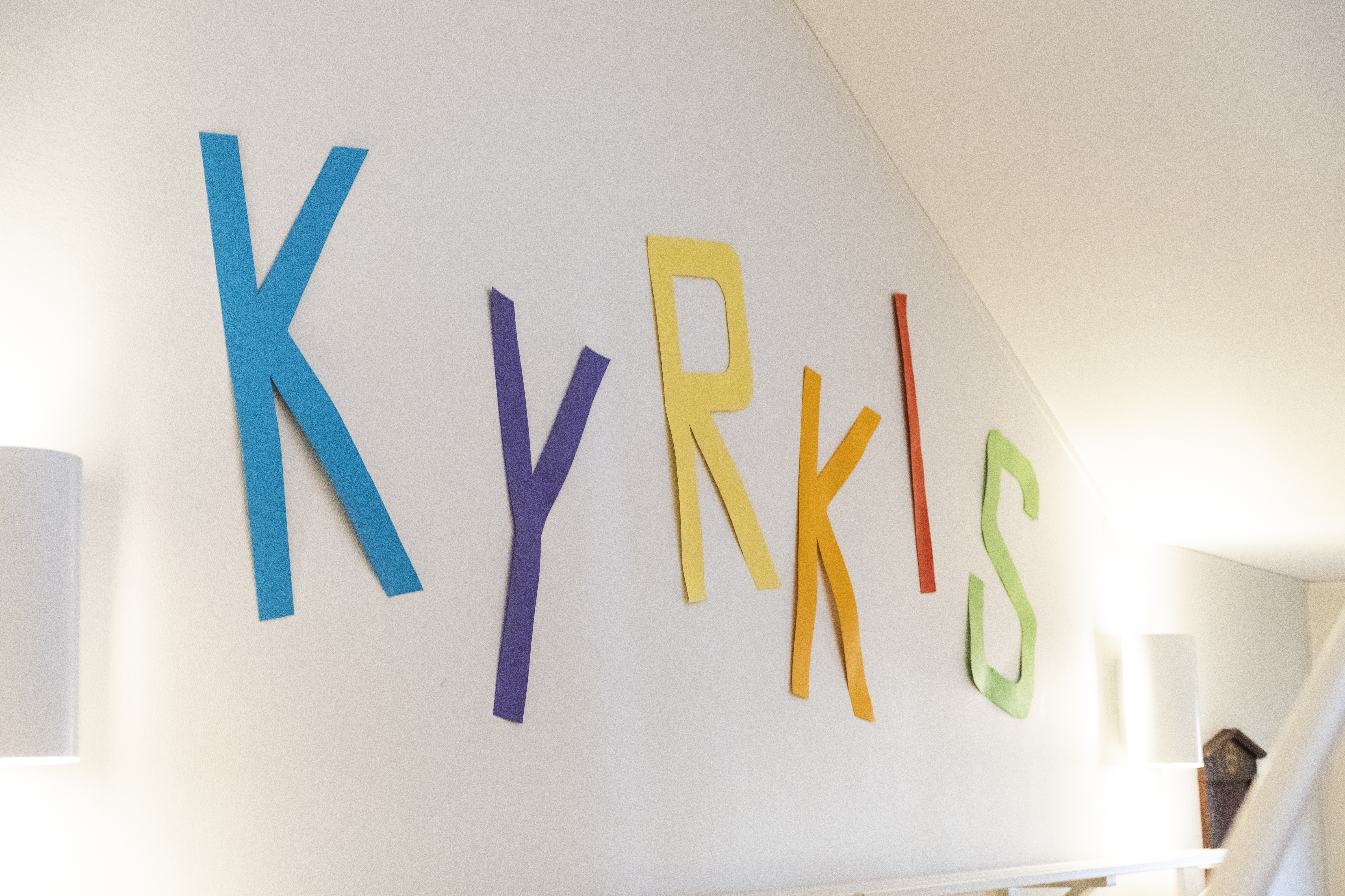 Ordet "KYRKIS" sitter upp på en vit vägg. Bokstäverna är urklippta ur färgat papper i blått, lila, gult, rött och grönt.