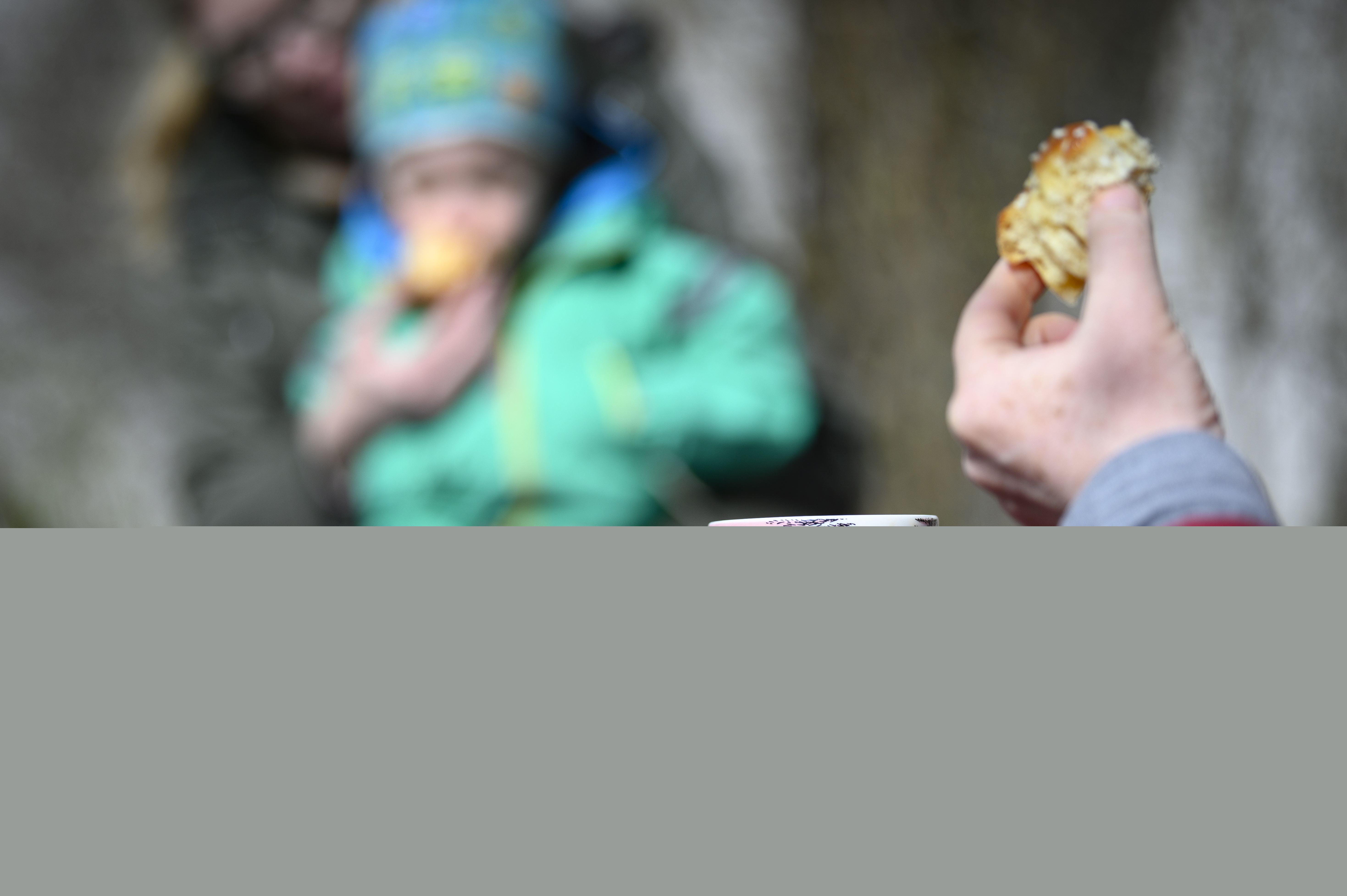 Någon sitter och fikar kaffe och bulle. I bakgrunden sitter ett barn i knät på en vuxen.