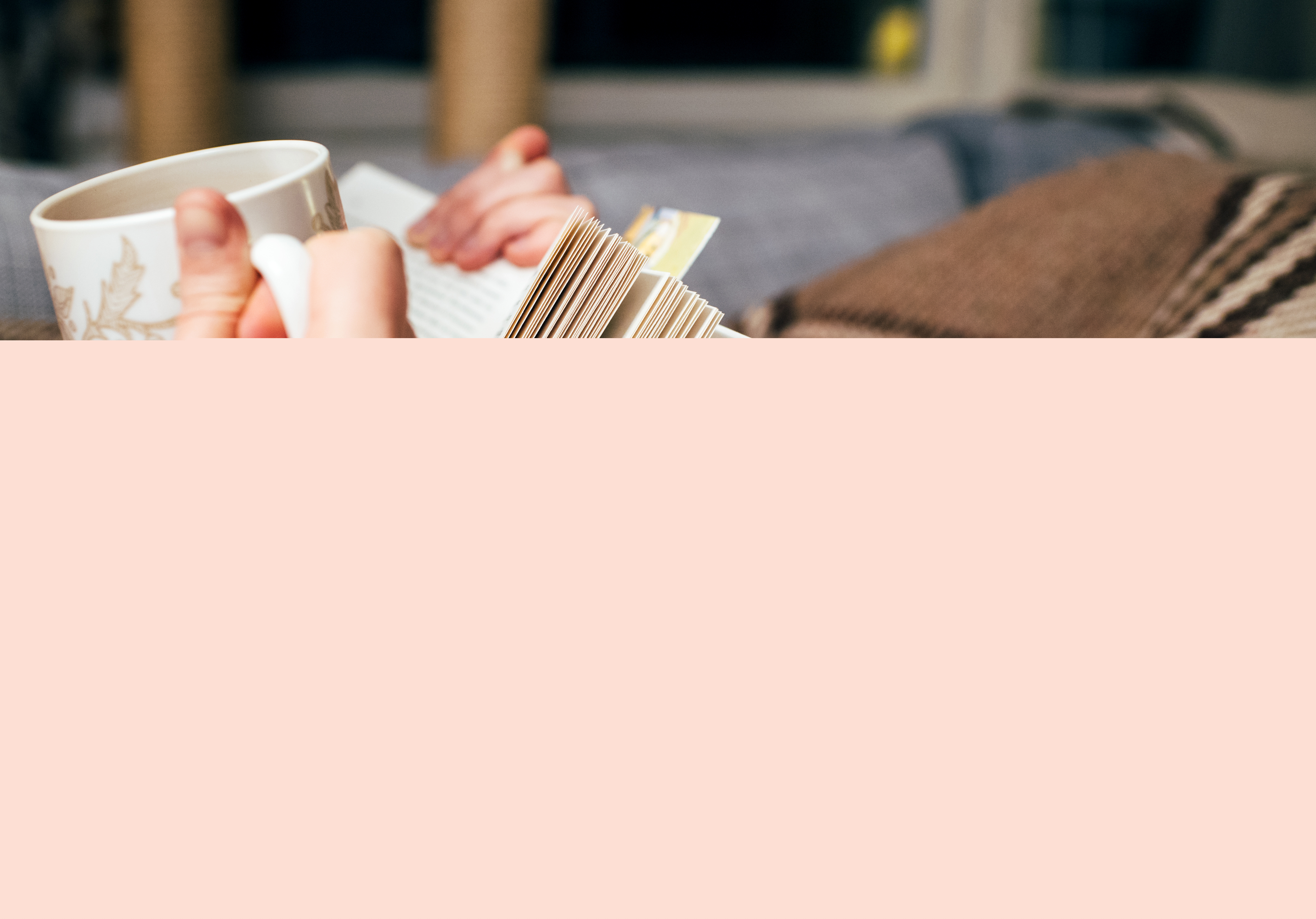 Närbild på någon som ligger under en filt och läser med kopp i handen.