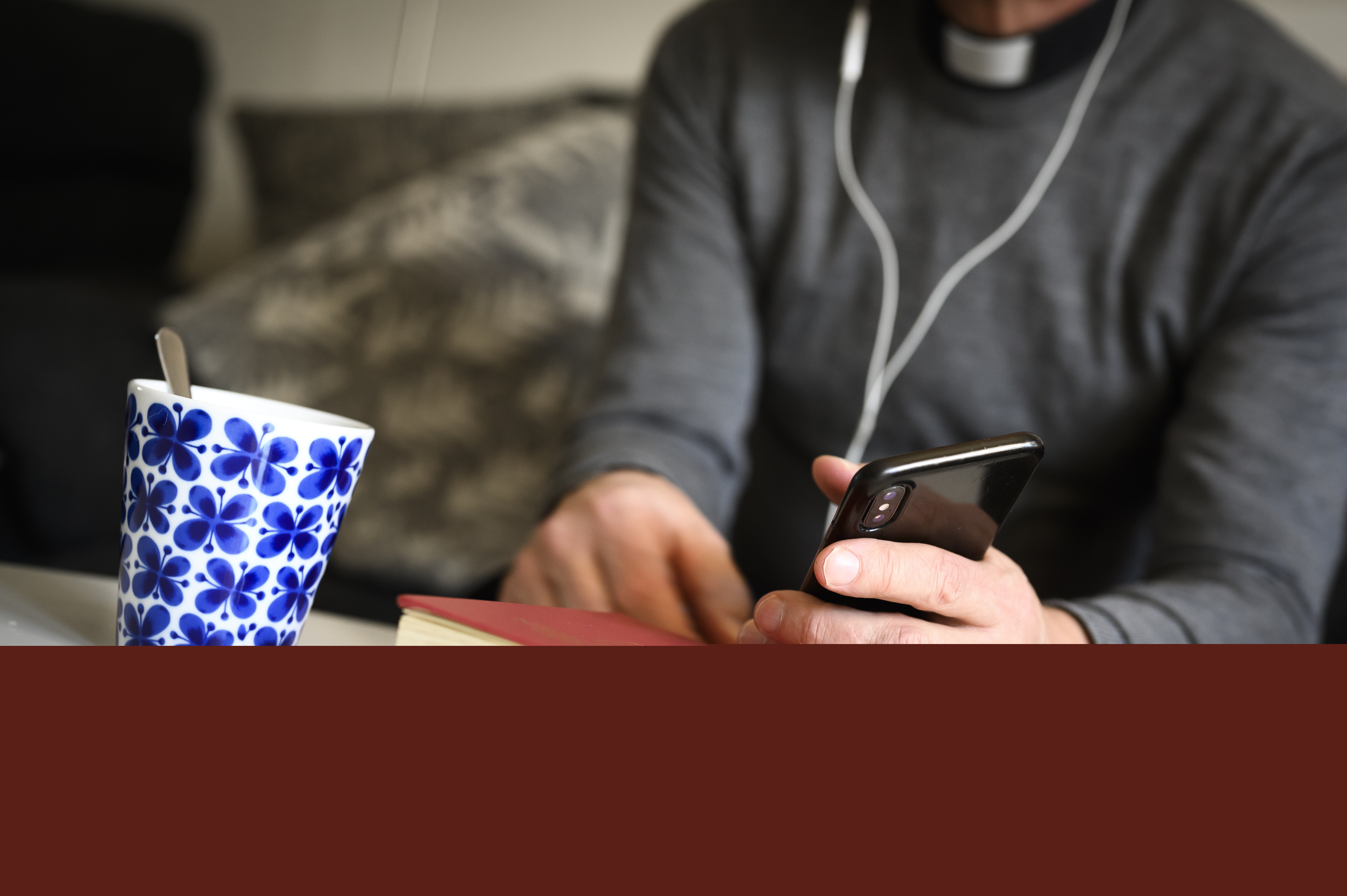 En präst sitter med hörlurar och pratar i mobiltelefon. En psalmbok ligger på bordet framför.