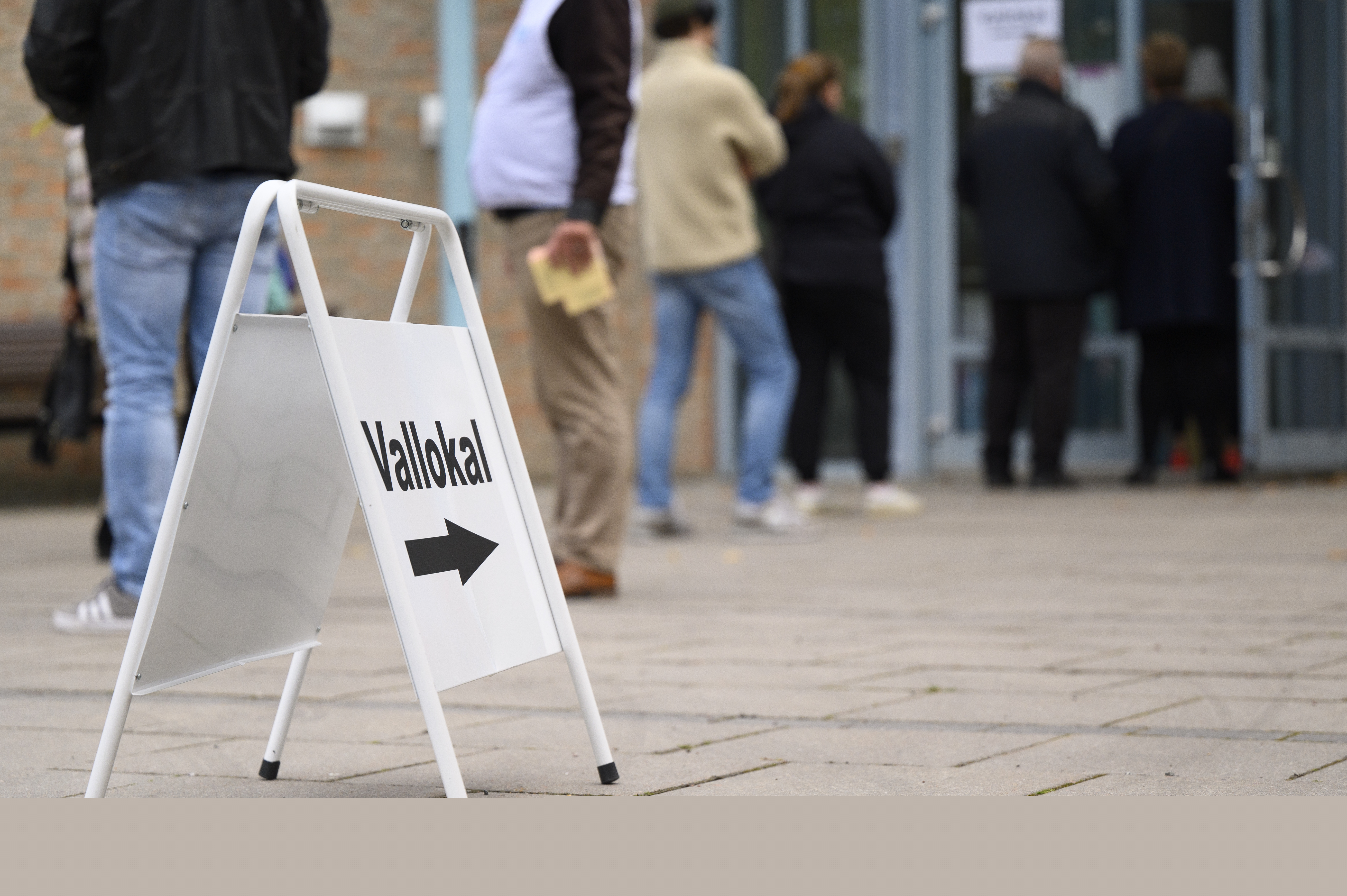En skylt med texten Vallokal står på marken. Människor köar utanför en öppen dörr i bakgrunden.