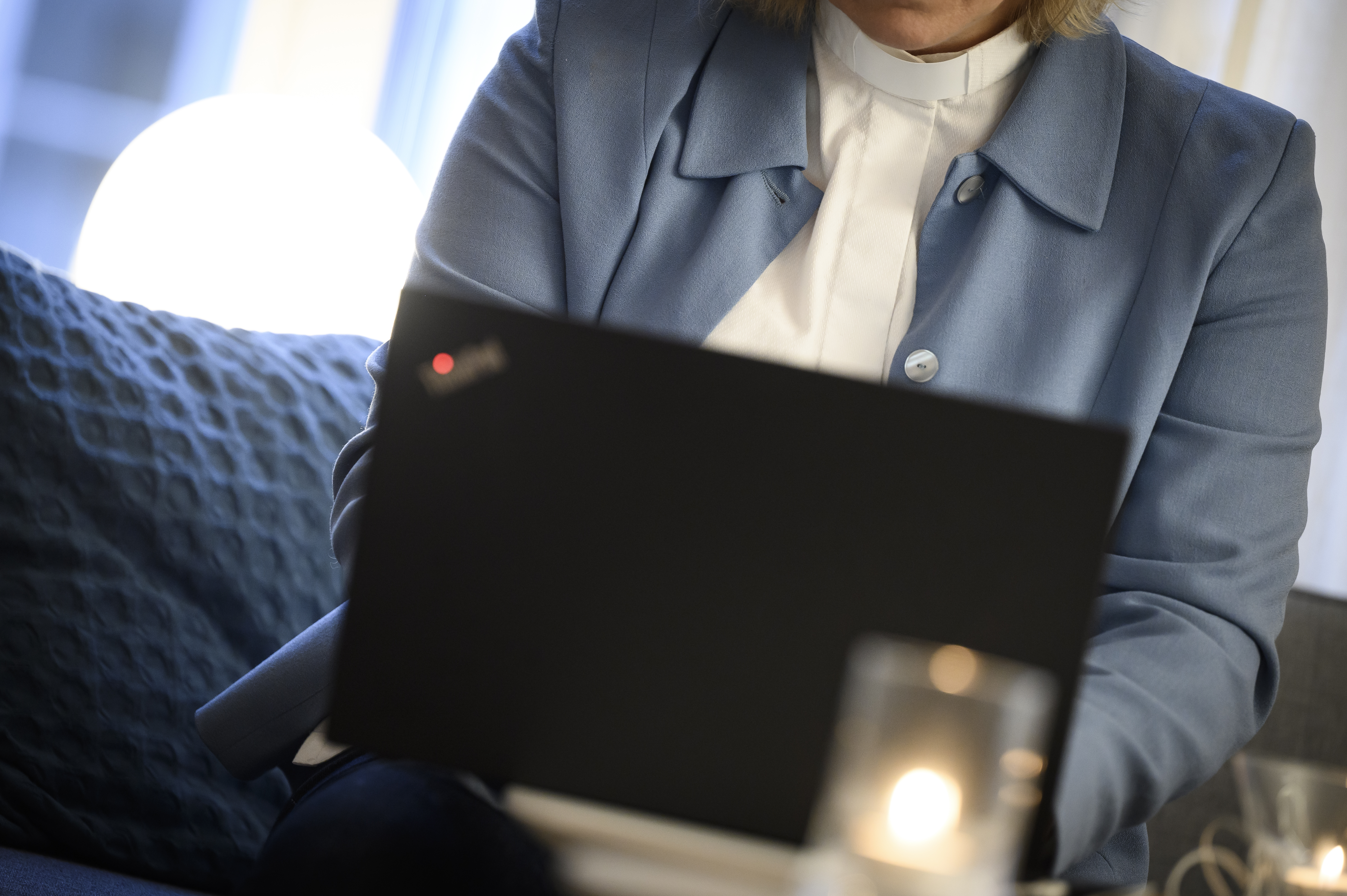 En kvinnlig präst i ljusblå kavaj sitter i en soffa med en laptop i knät och en mobiltelefon i handen.