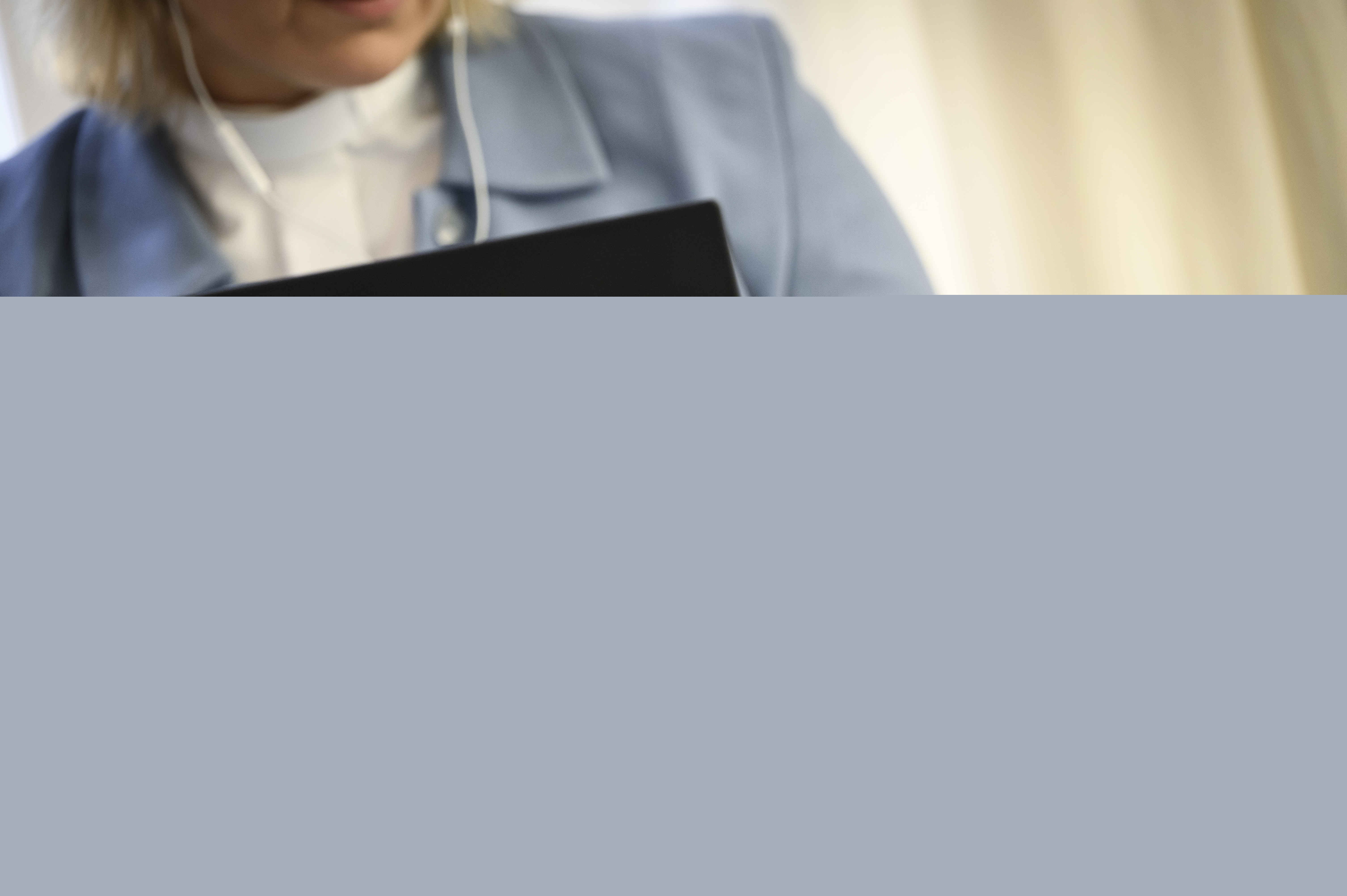 En kvinnlig präst i ljusblå kavaj sitter i en soffa med en laptop i knät och en mobiltelefon i handen.