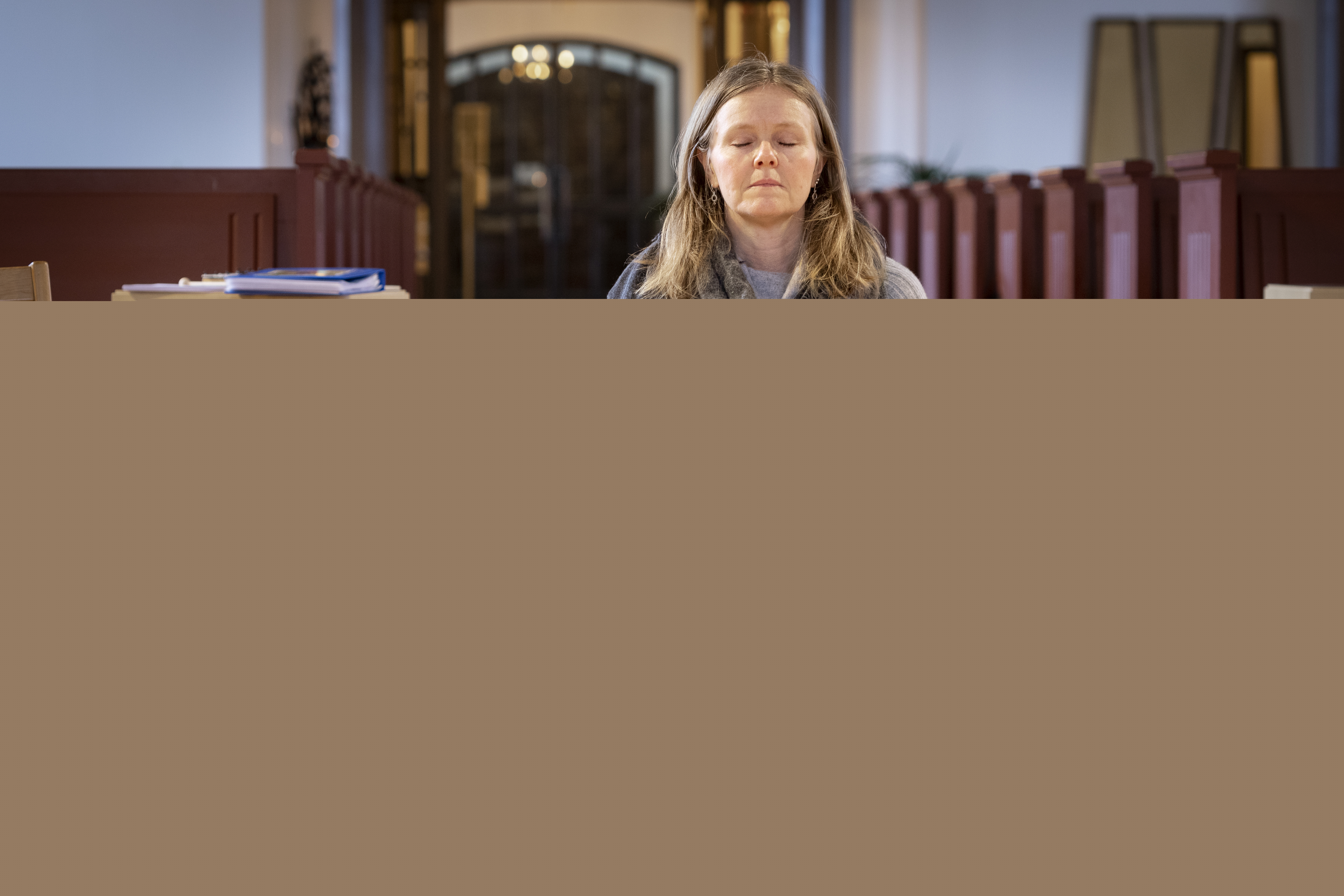 En kvinna sitter på en bönepall på en kyrkas golv, med knäppta händer och slutna ögon.