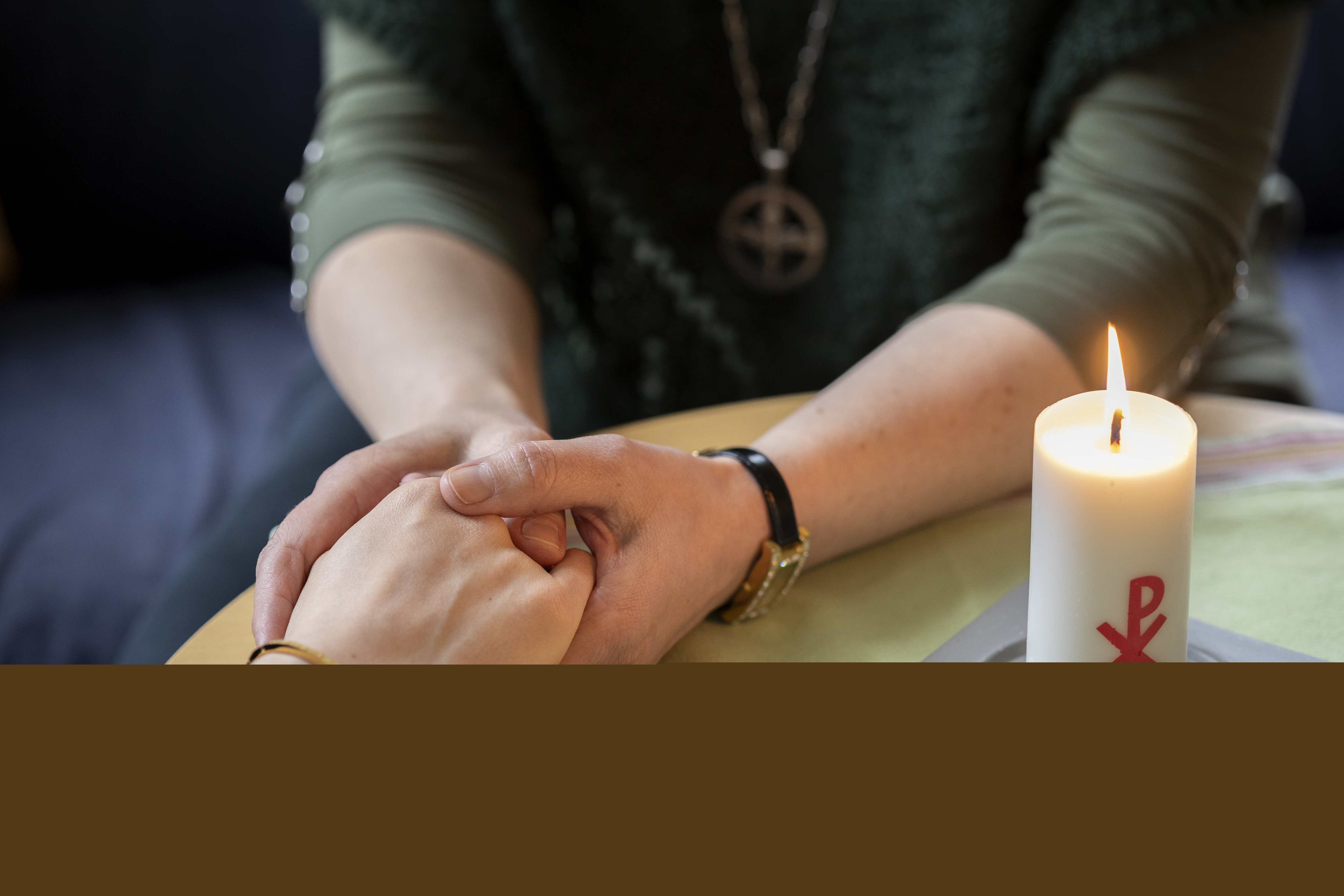 En diakon håller en annans hand i sina händer. Ett stearinljus med kristusmonogram brinner på bordet mellan dem.