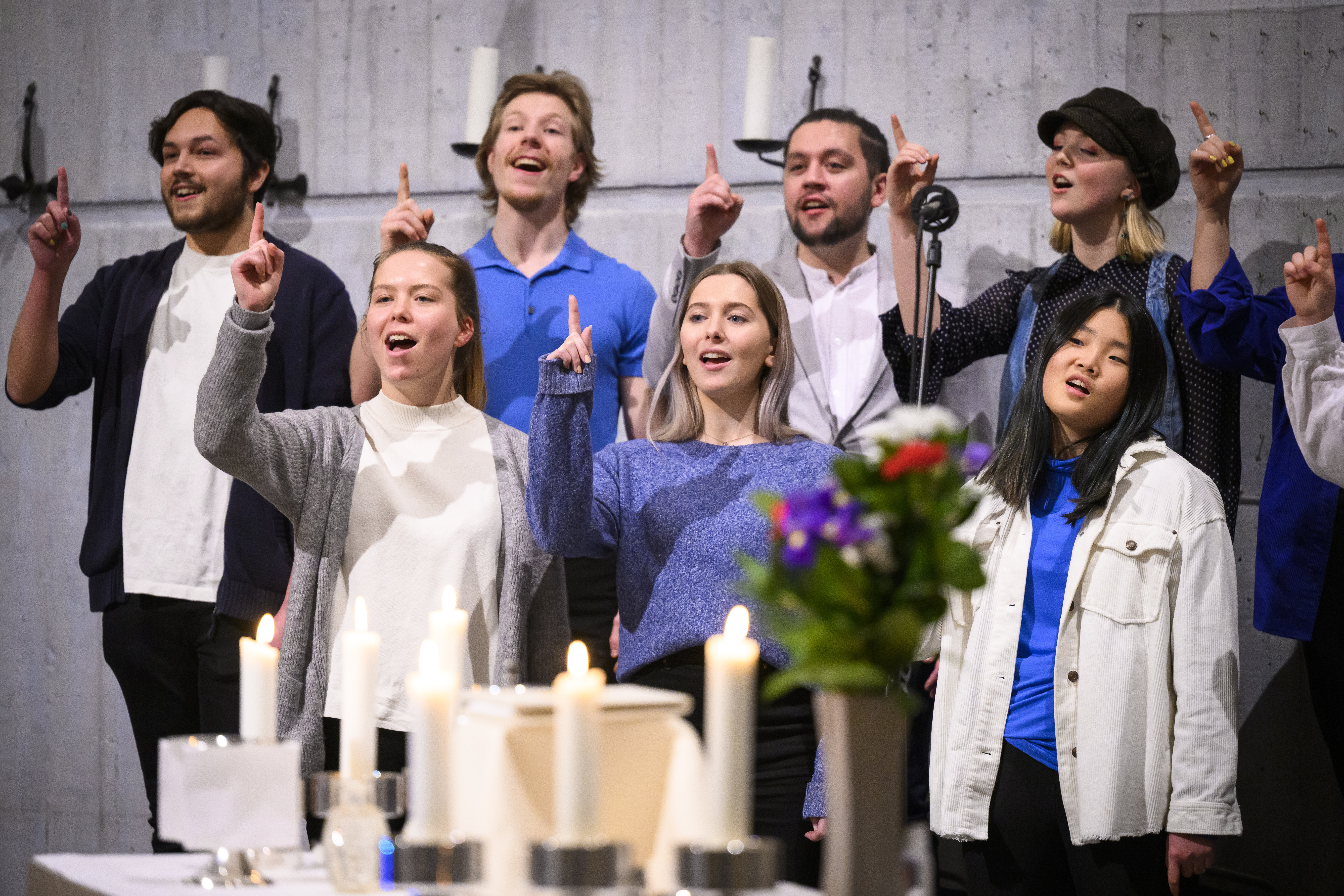 En blandad ungdomskör sjunger i en kyrka. De håller upp sina pekfingrar.