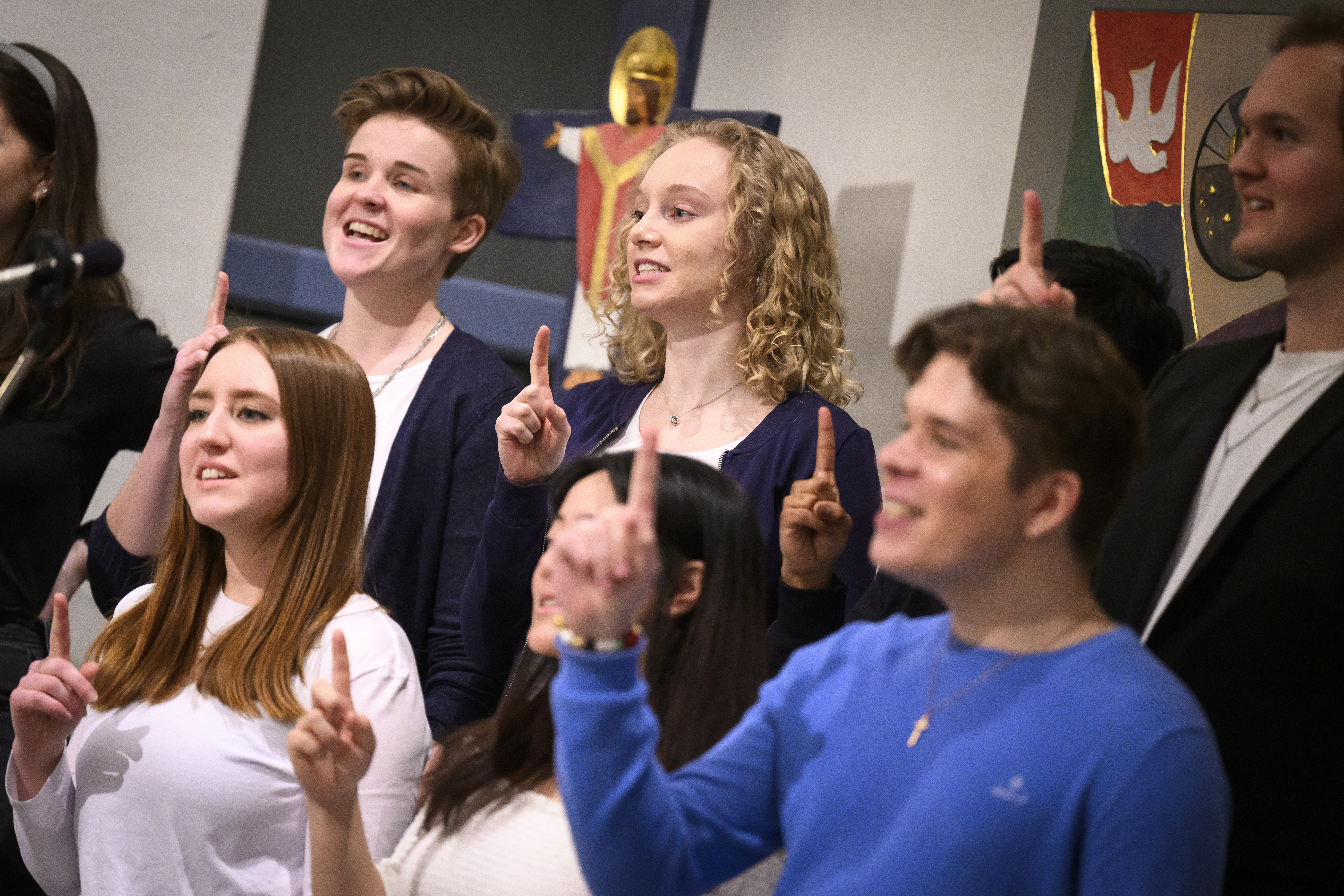 En blandad ungdomskör sjunger i en kyrka. De håller upp sina pekfingrar.