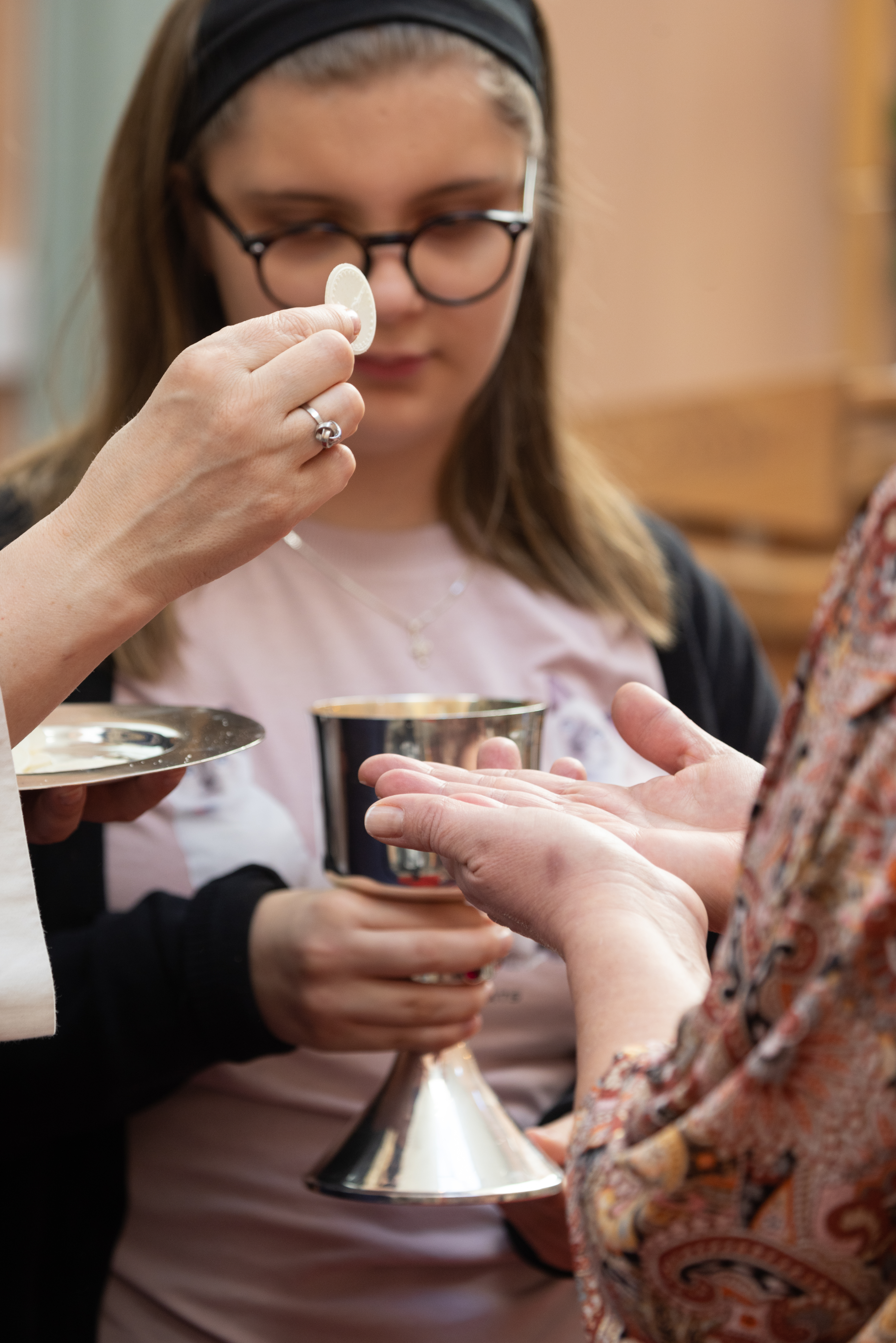 En hand håller upp en oblat mot någons utsträckta händer. En flicka håller en silverbägare.