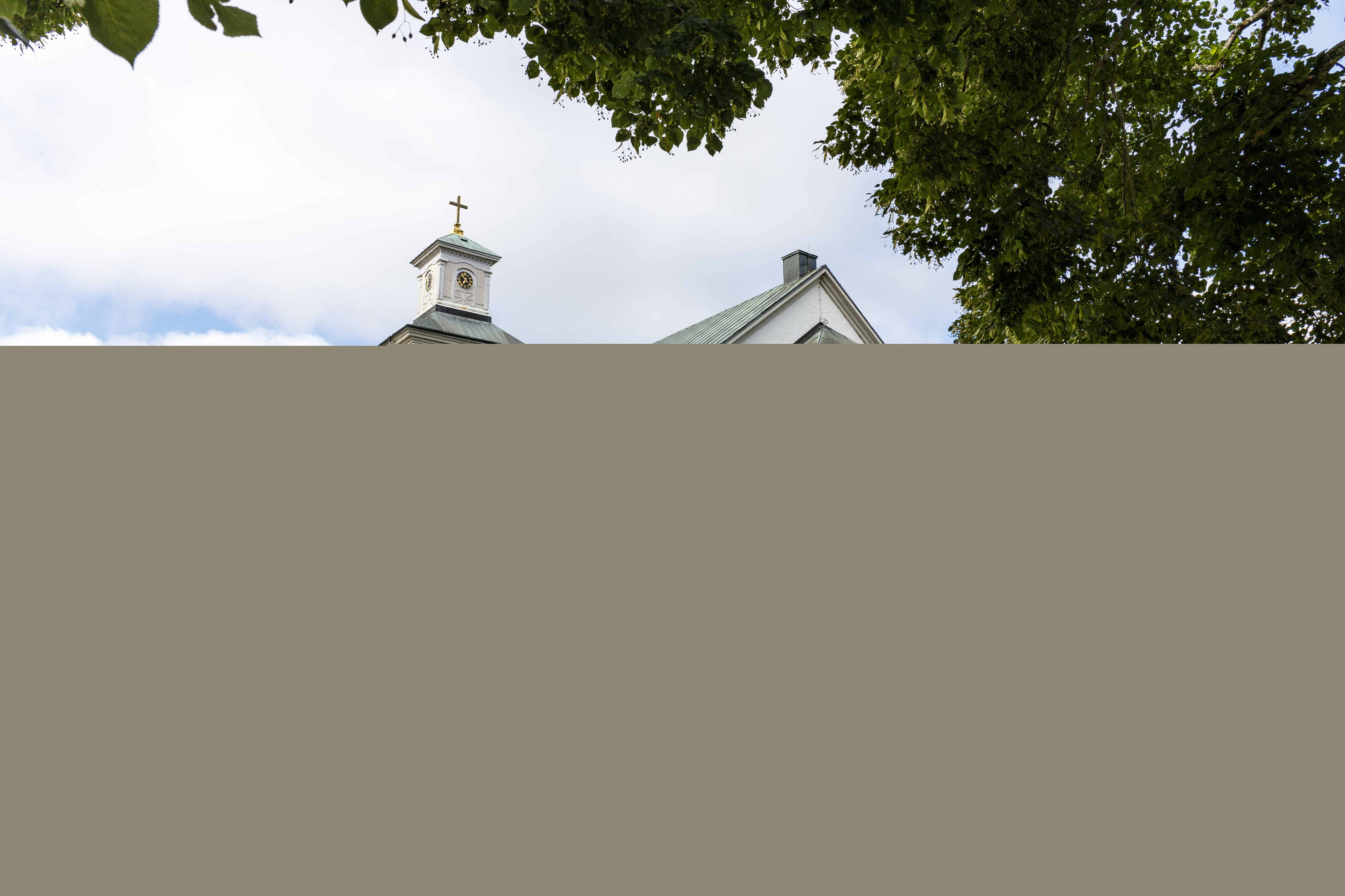 En vitputsad stenkyrka med ärgat tak. Gravstenar och grönskande träd i omgivningen.