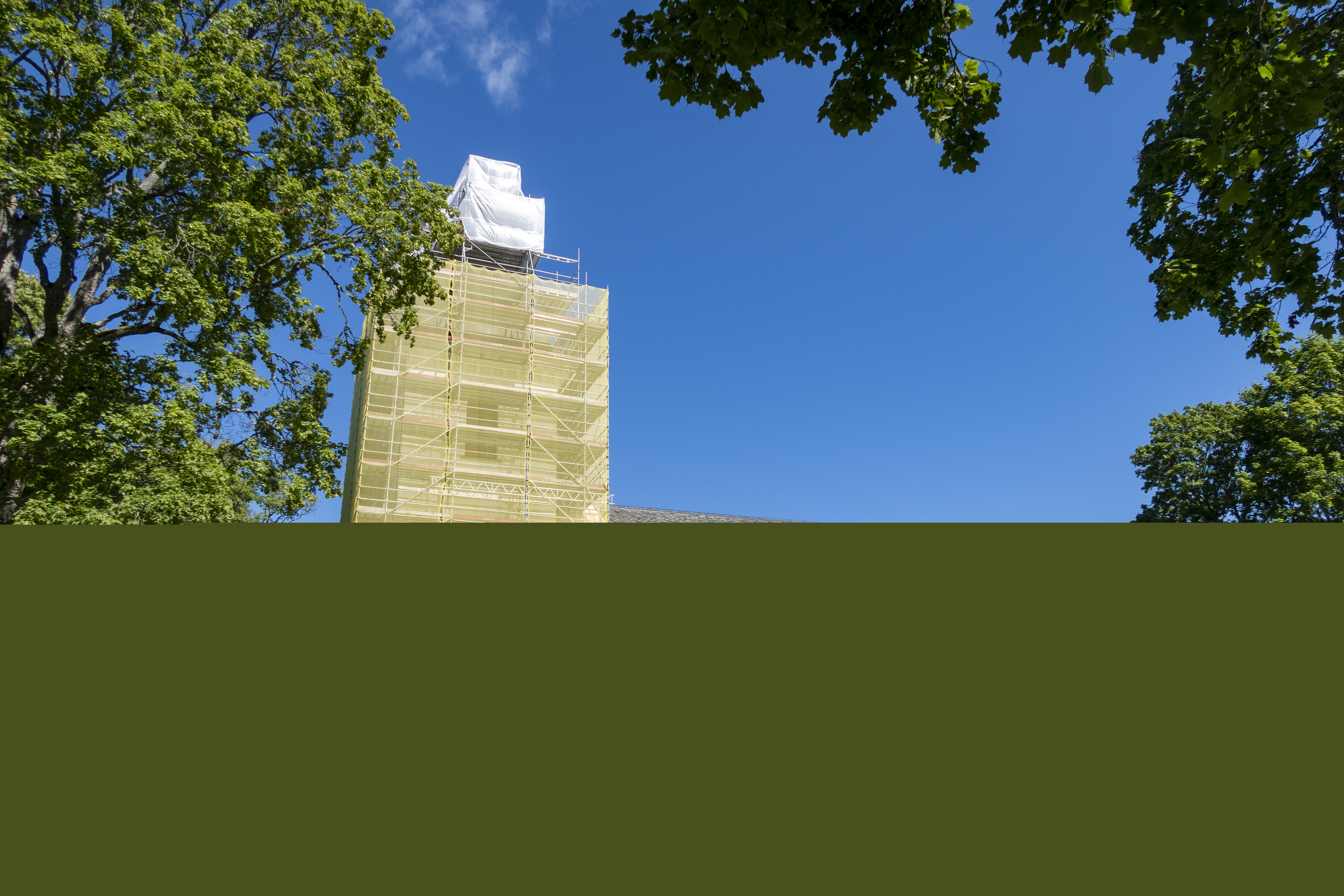 Byggnadsställningar runt ett kyrktorn. En kyrkogård framför den vita kyrkan.