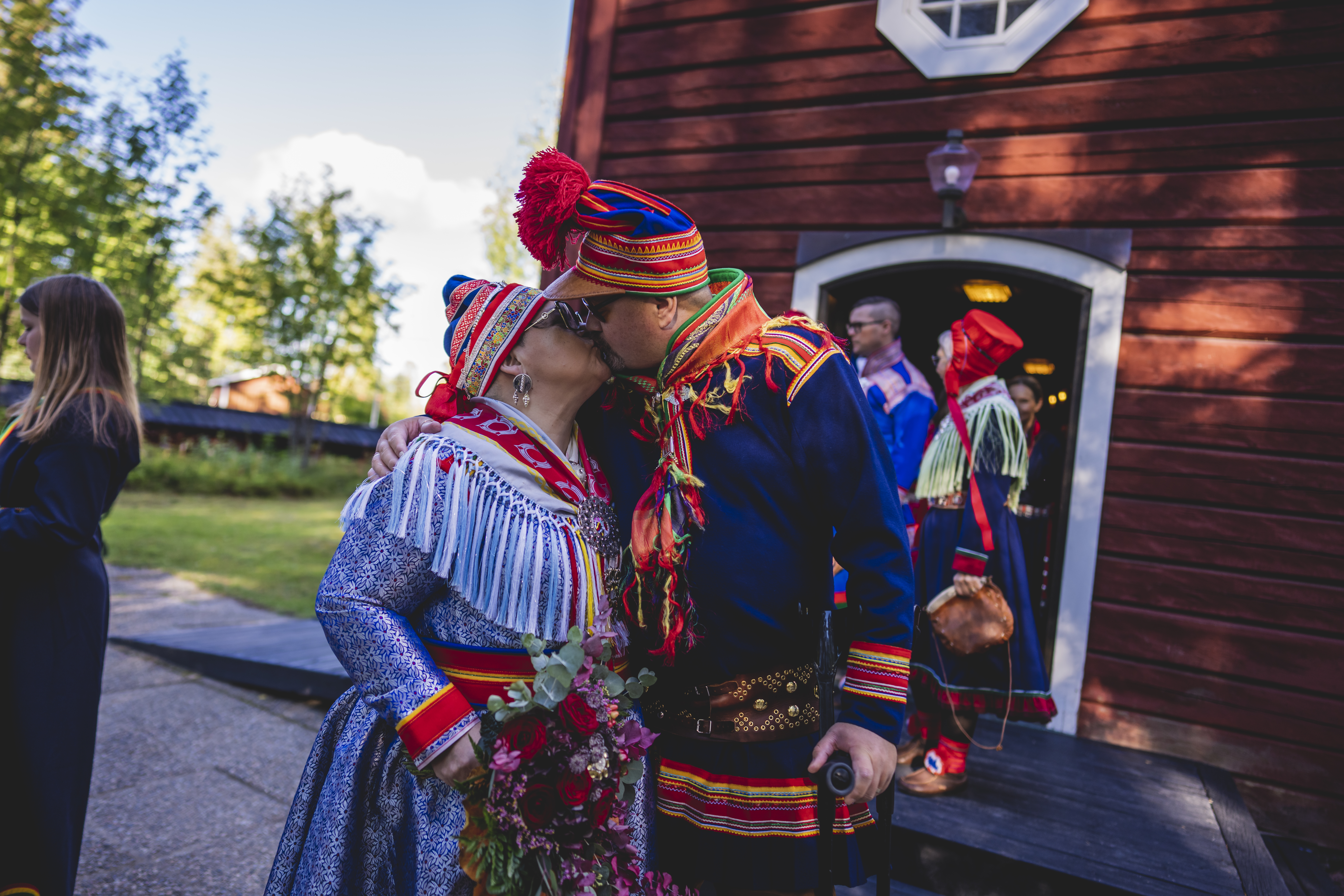 En man och en kvinna i samiska folkdräkter kysser varandra utanför en röd träkyrka. Fler är på väg ut ur kyrkan.