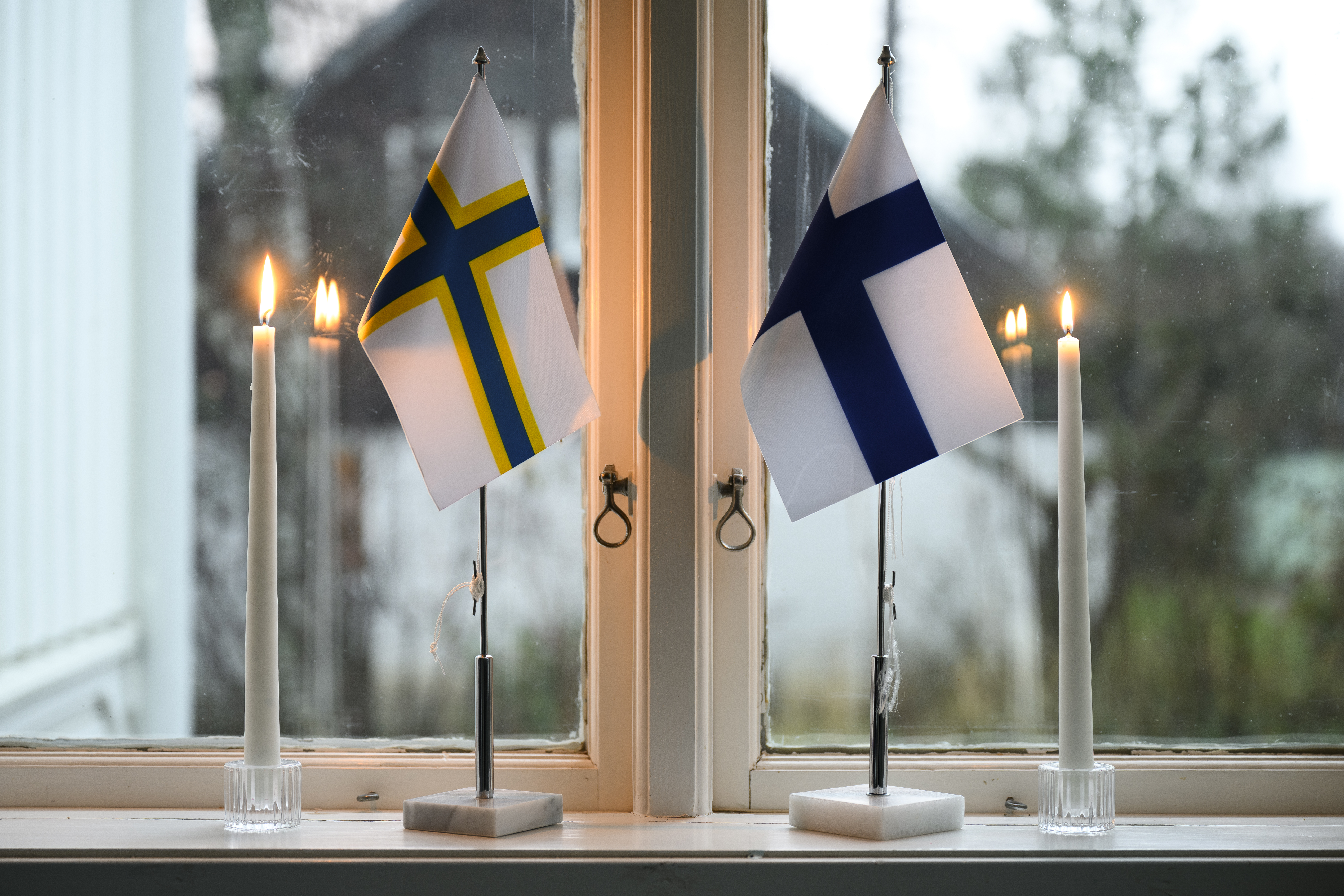 En finsk och en sverigefinsk bordsflagga och två brinnande stearinljus står i ett fönster.