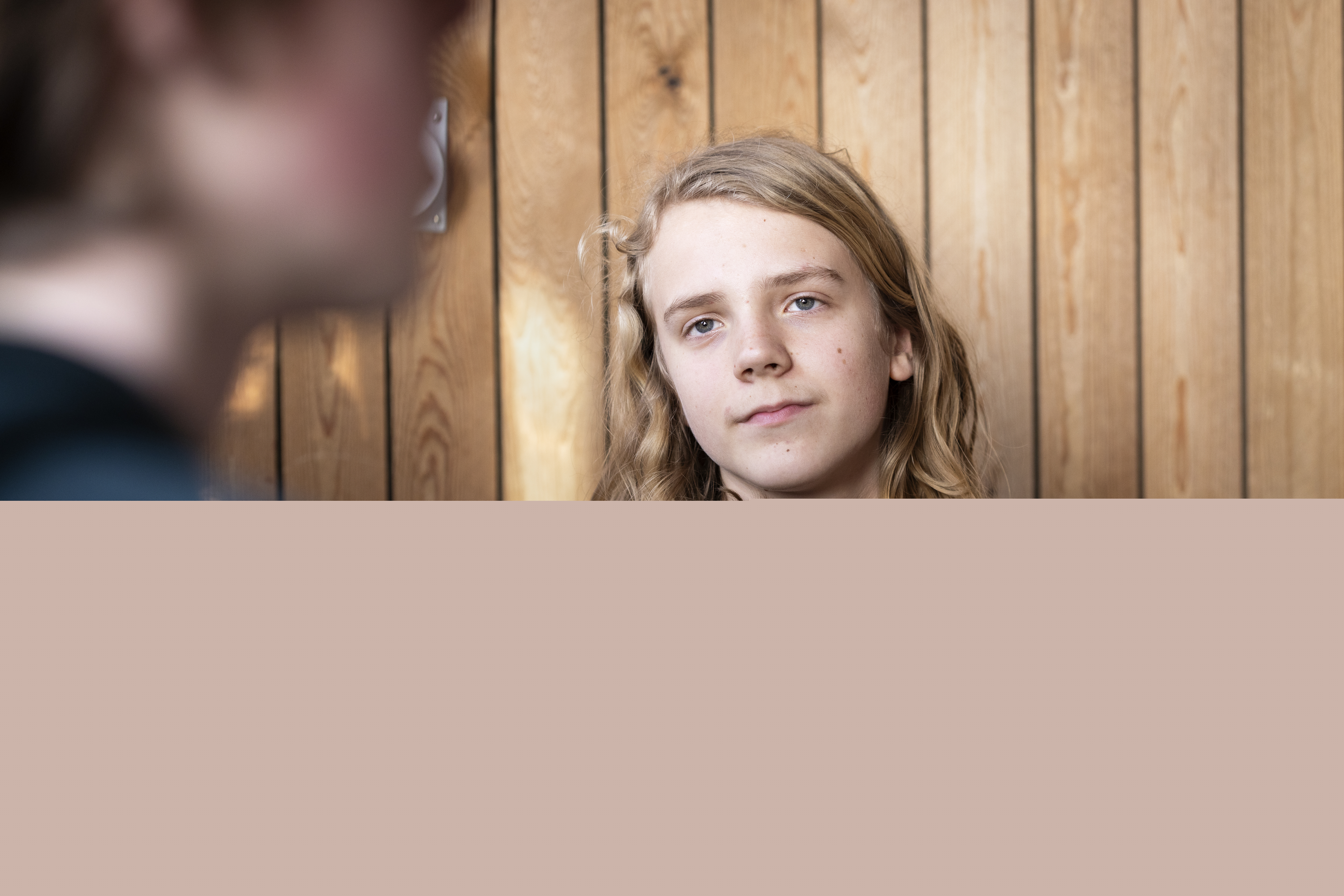 En ung kille med långt hår sitter i en soffa. Någon syns suddigt i förgrunden.