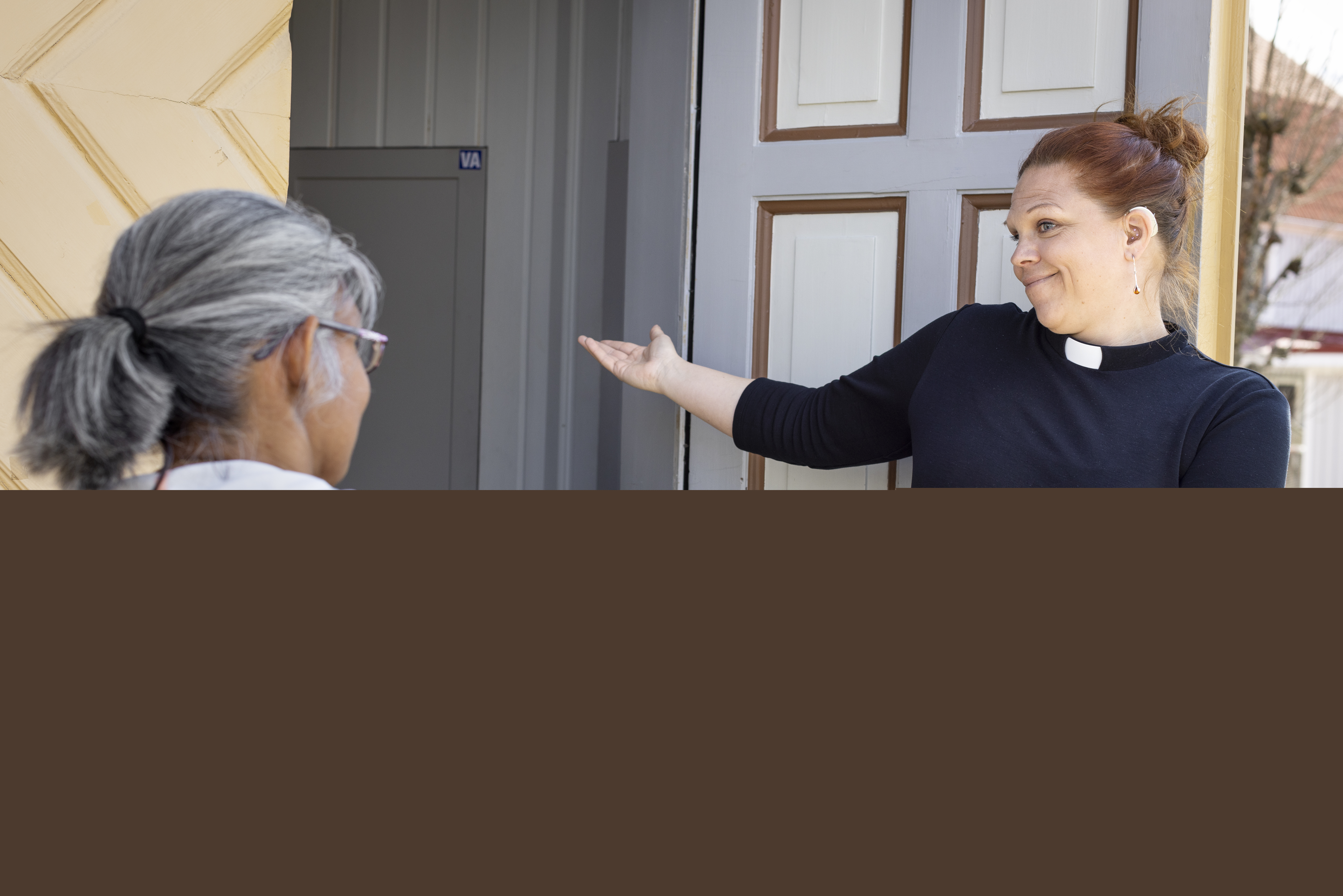 En kvinnlig präst visar vägen in i kyrkan med armarna åt en kvinna.