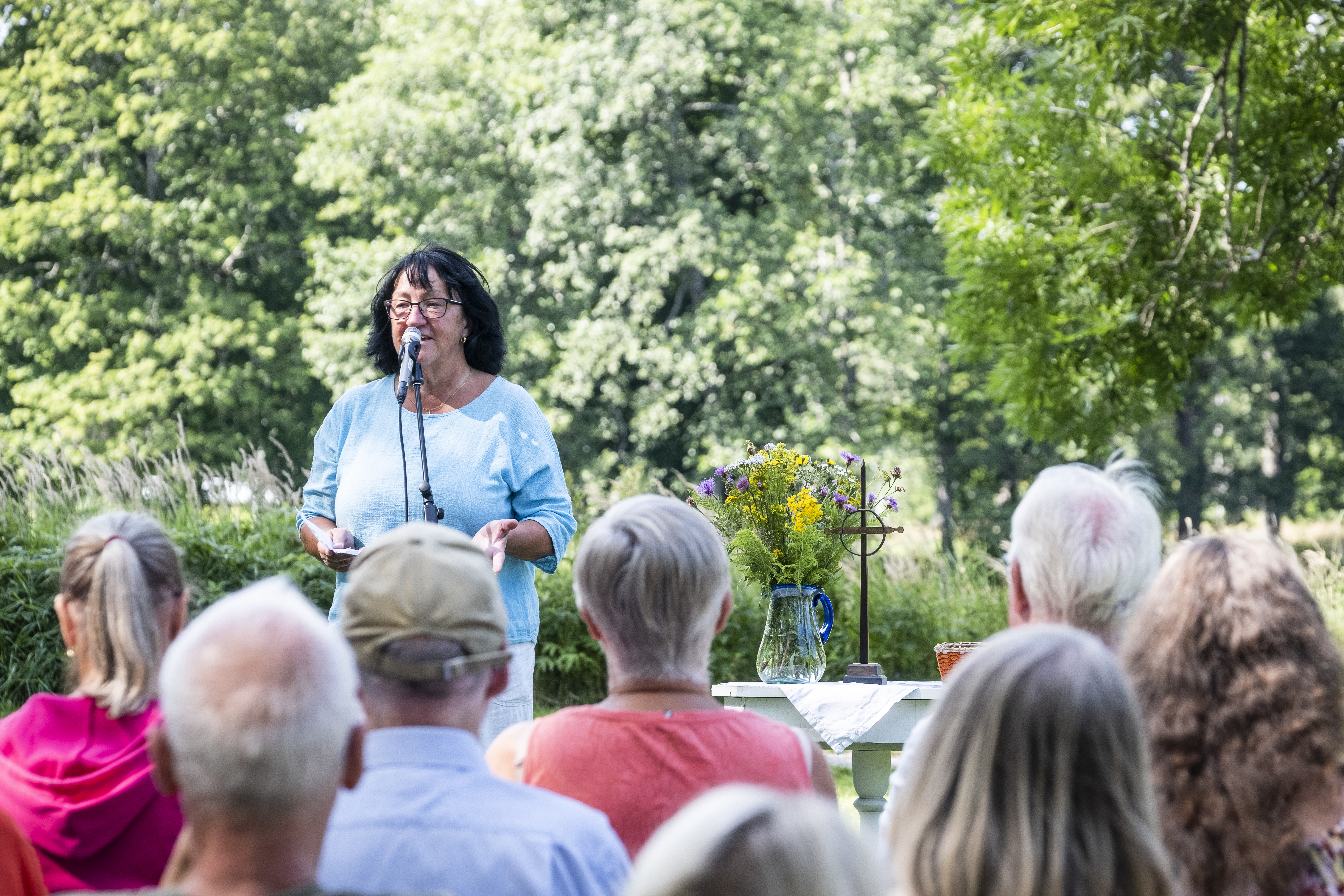 En kvinna pratar i en mikrofon utomhus. Åskådare i förgrunden och blomstrande natur i bakgrunden.