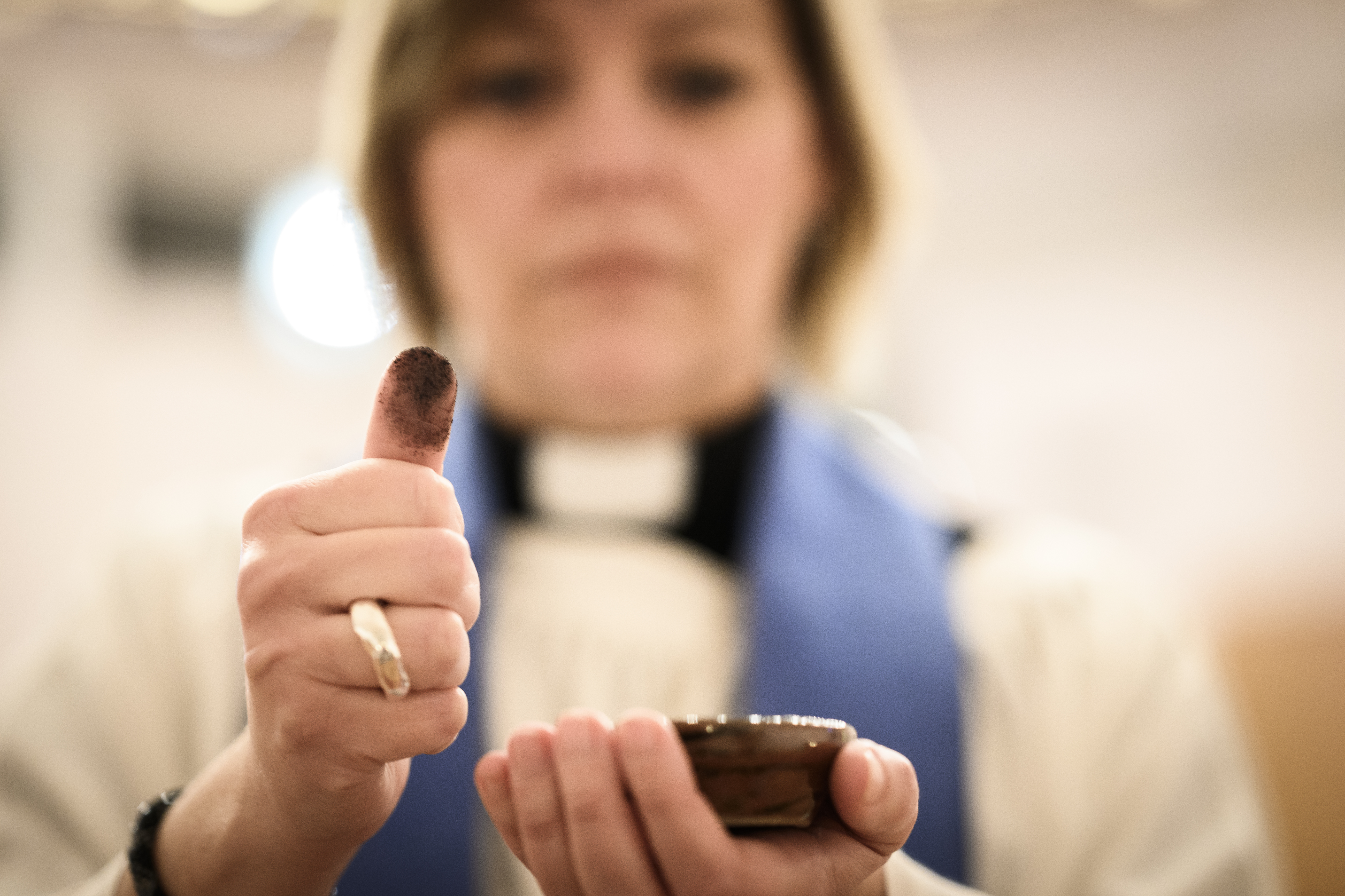 En kvinnlig präst håller upp sin tumme, svart av aska, och en lite skål mot kameran.