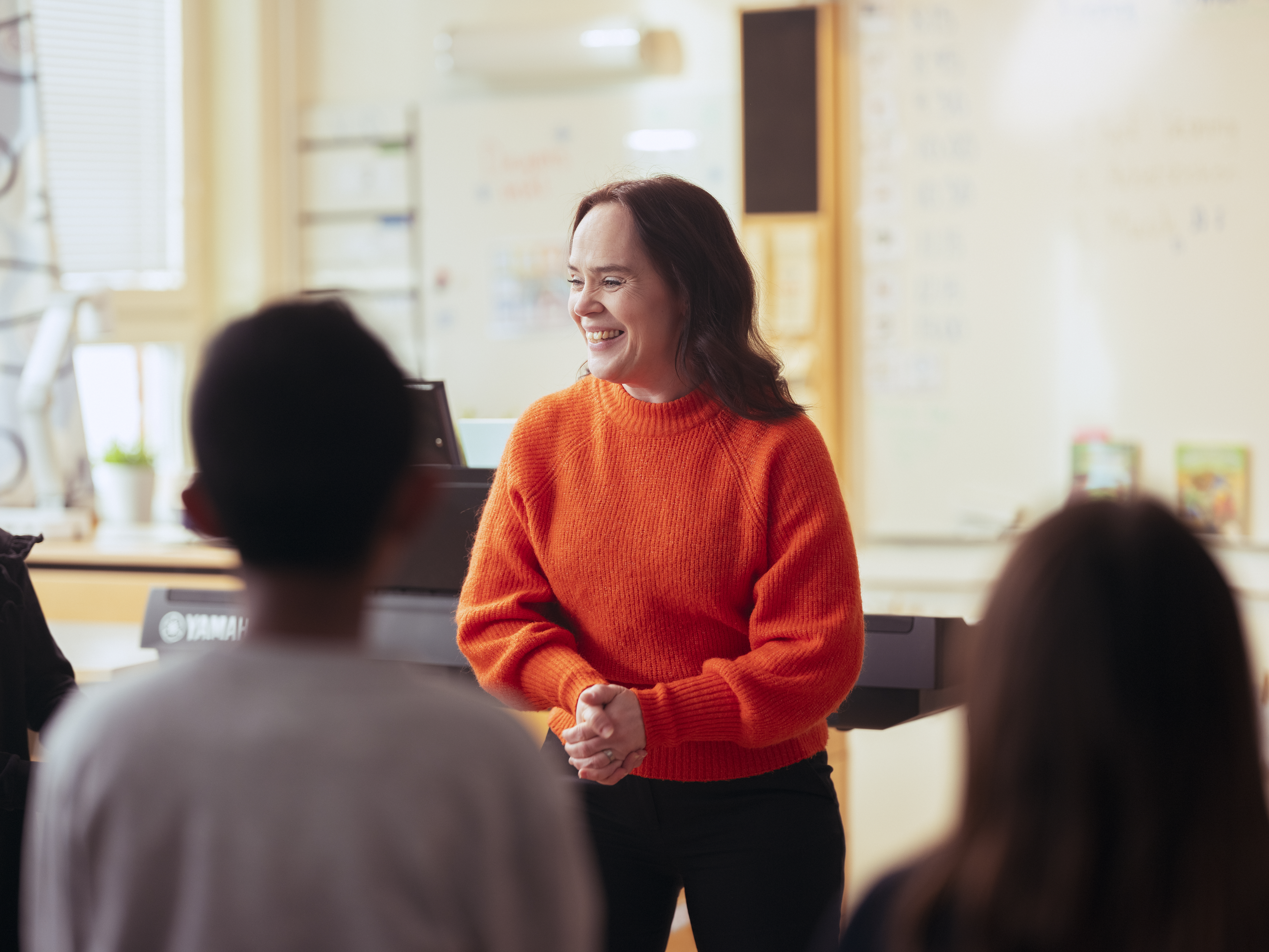 En kvinna i orange stickad tröja står och ler framför några barn i ett klassrum.