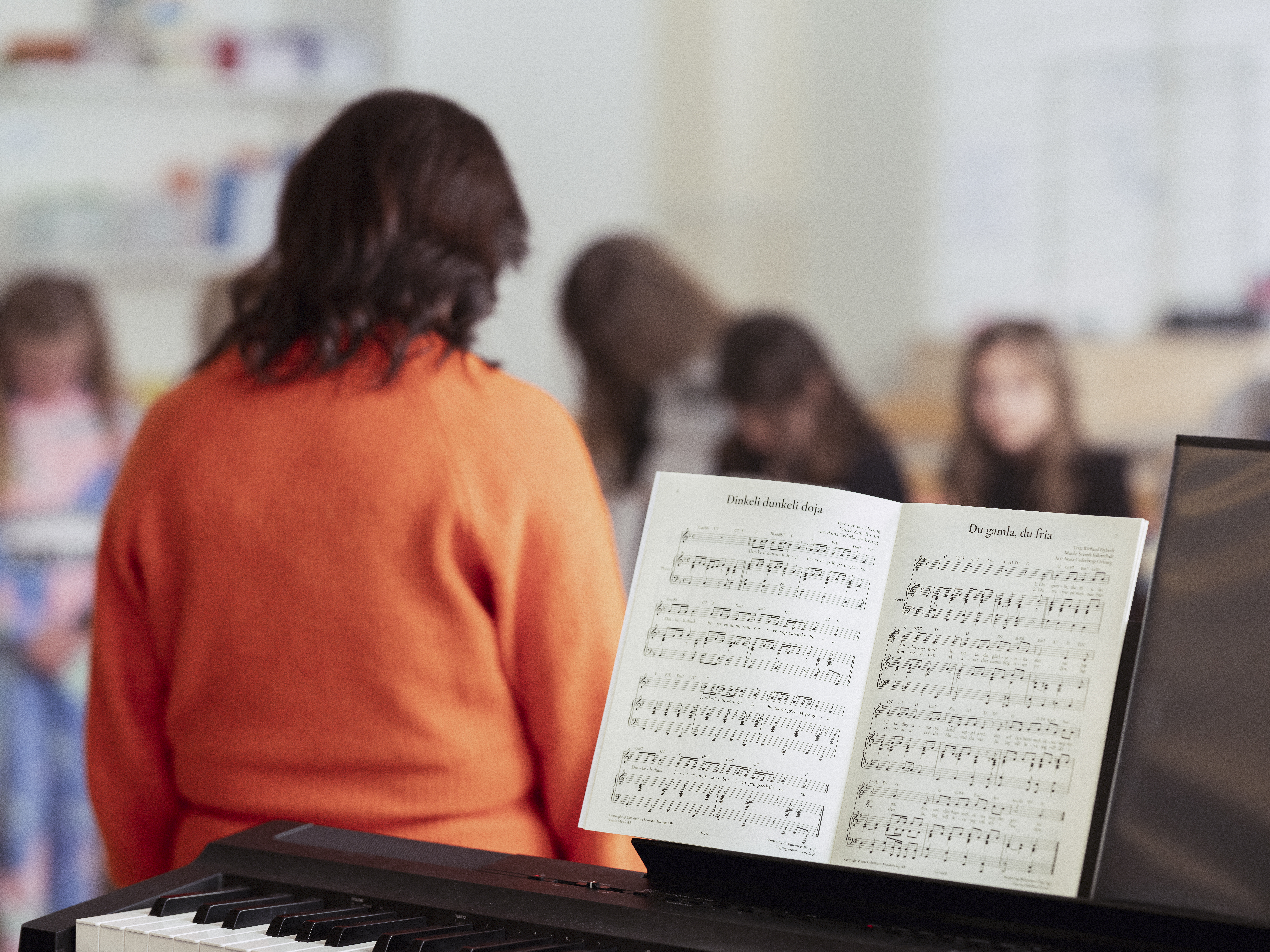 En kvinnlig körledare står framför en barnkör. En keyboard med ett nothäfte står i förgrunden.