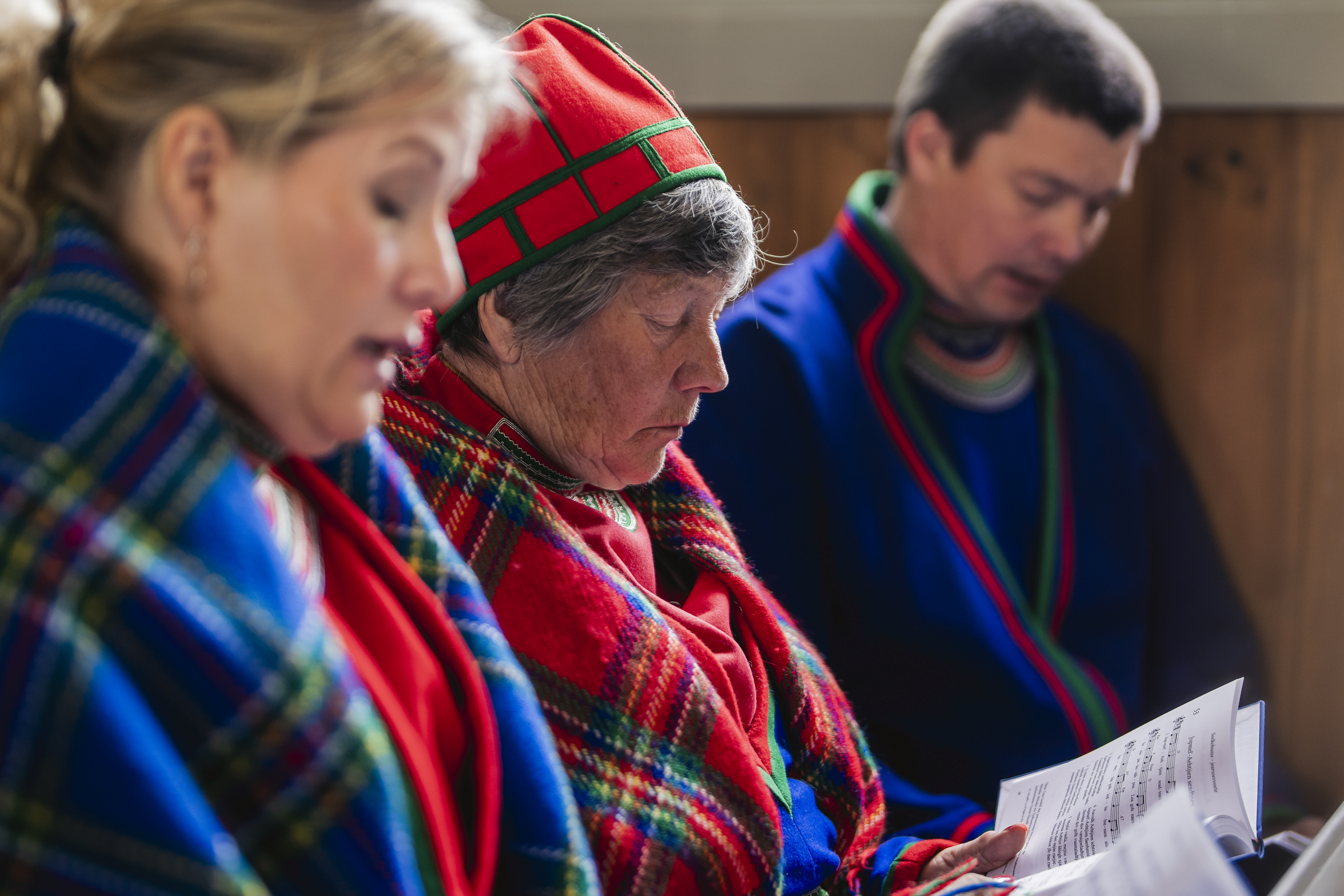 Tre personer i samiska kläder sjunger i kyrkbänken.