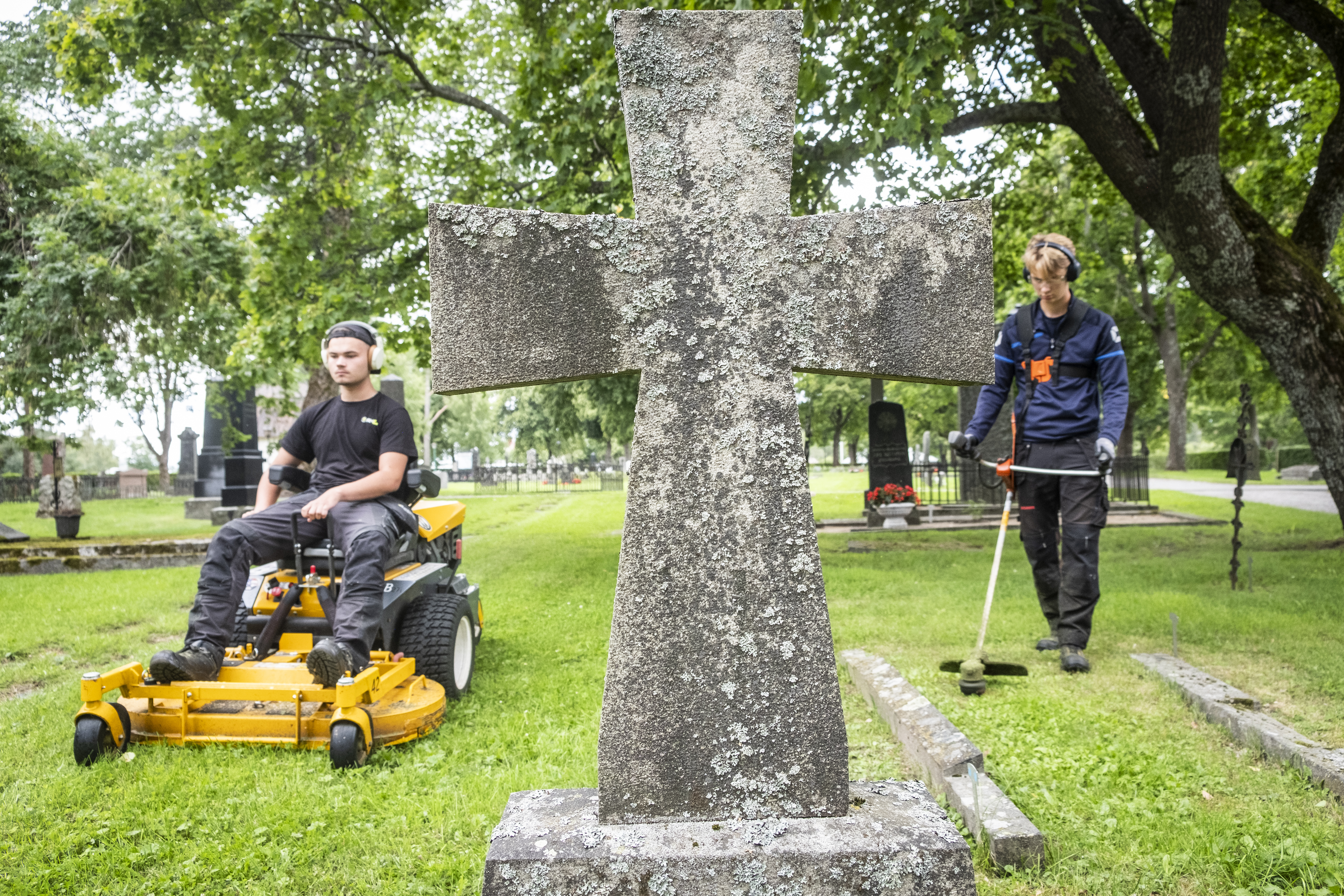 Två unga män klipper och trimmar gräsmattan på en kyrkogård. En av dem kör en åkgräsklippare. En gravsten i form av ett kors i förgrunden.