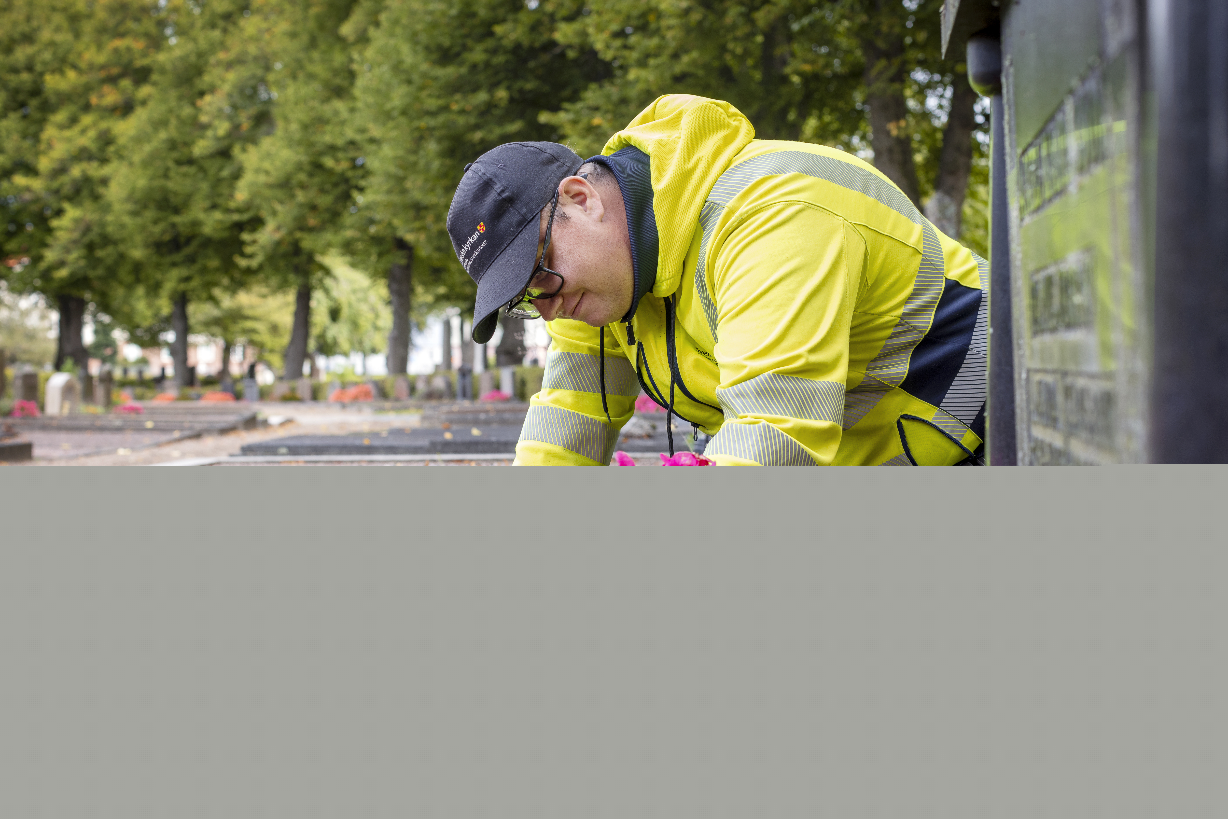 En kyrkogårdsarbetare i gula varselkläder fixar blommorna på en gravplats.