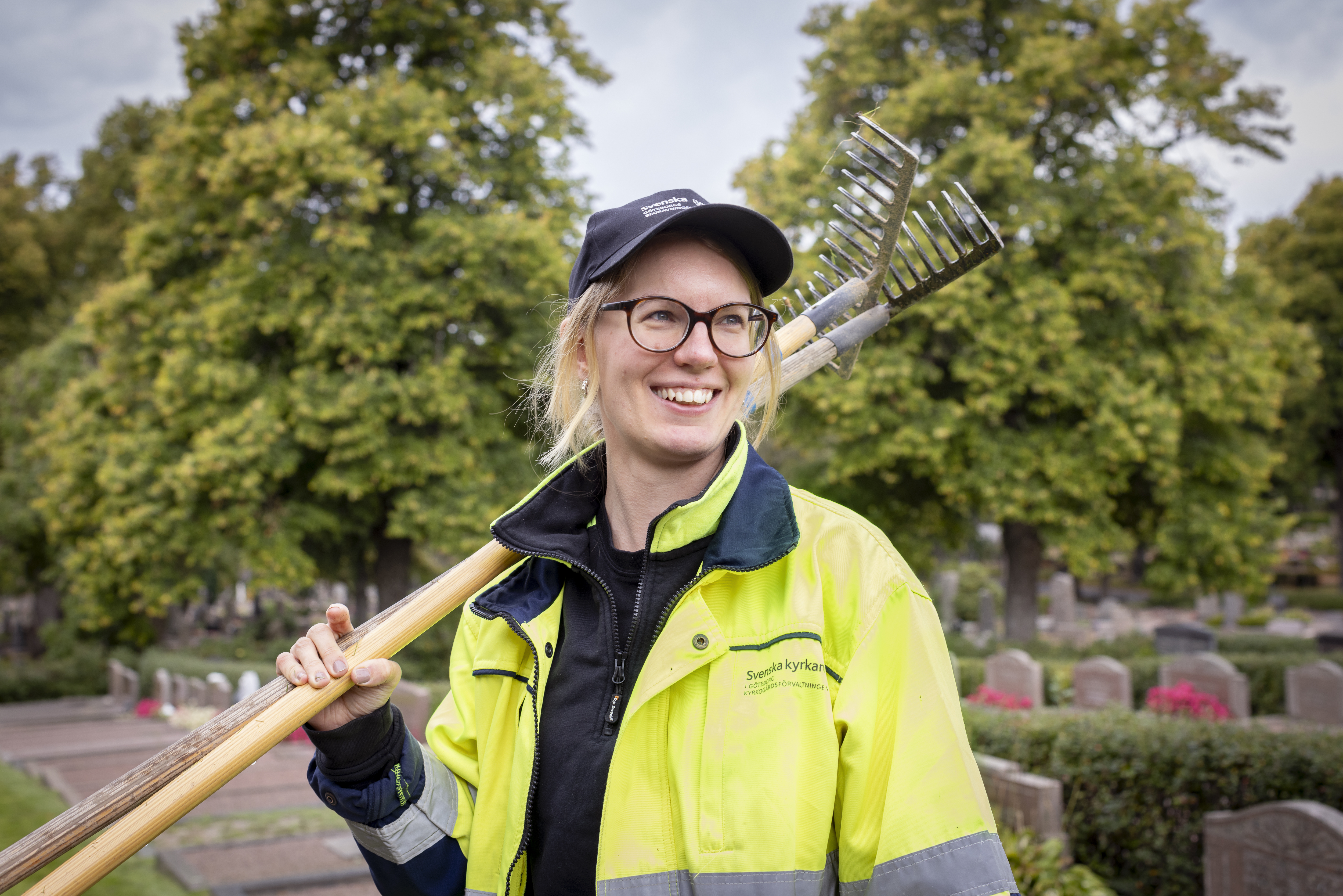 En kvinna i gula varselkläder och keps står med två krattor över axeln på en kyrkogård. Hon ler stort.