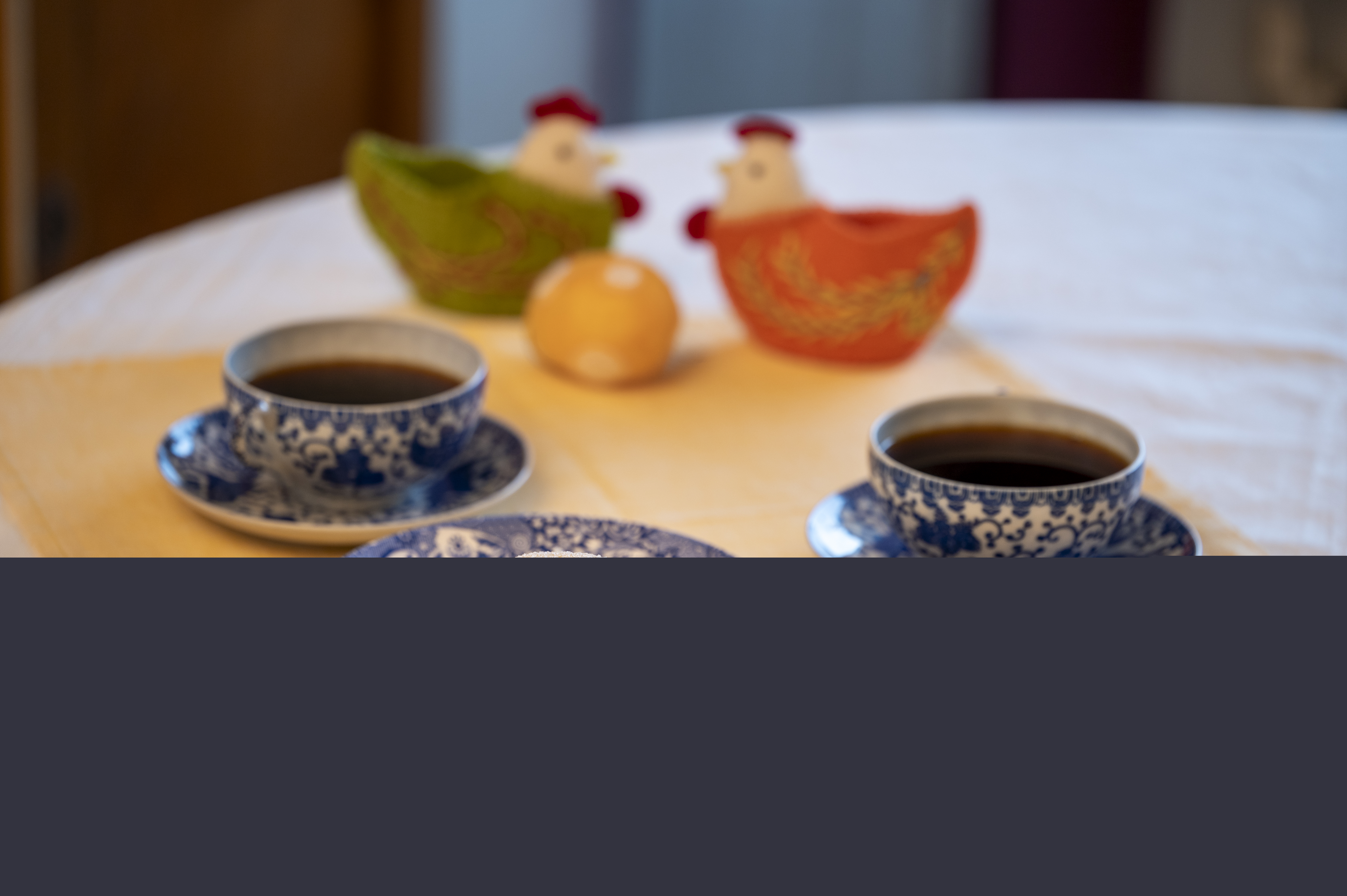 Ett kakfat, två kaffekoppar på fat och två påskhönor med ägg står uppdukade på en gul duk.