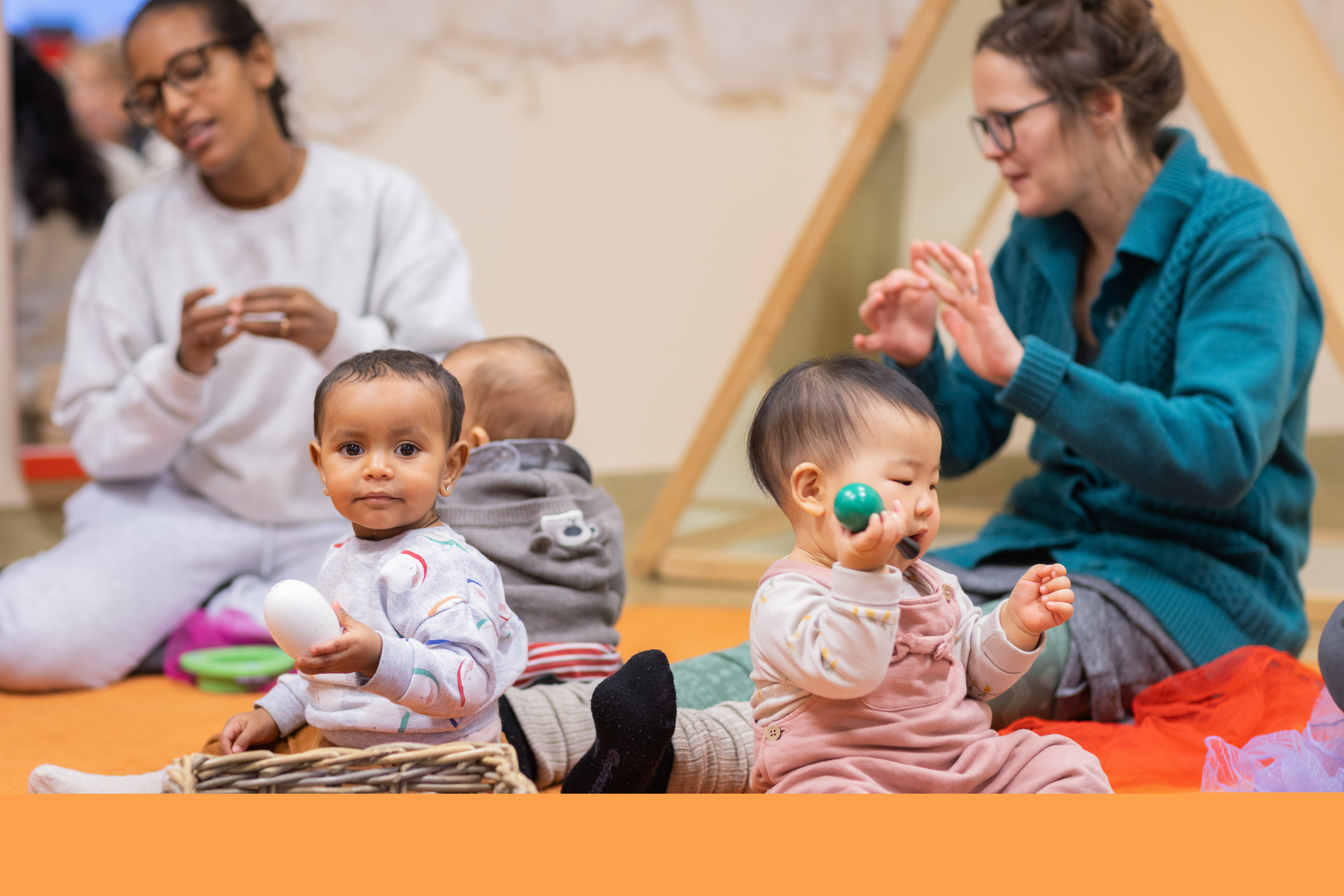 Tre bebisar sitter på golvet. Runt om sitter deras mammor och sjunger.