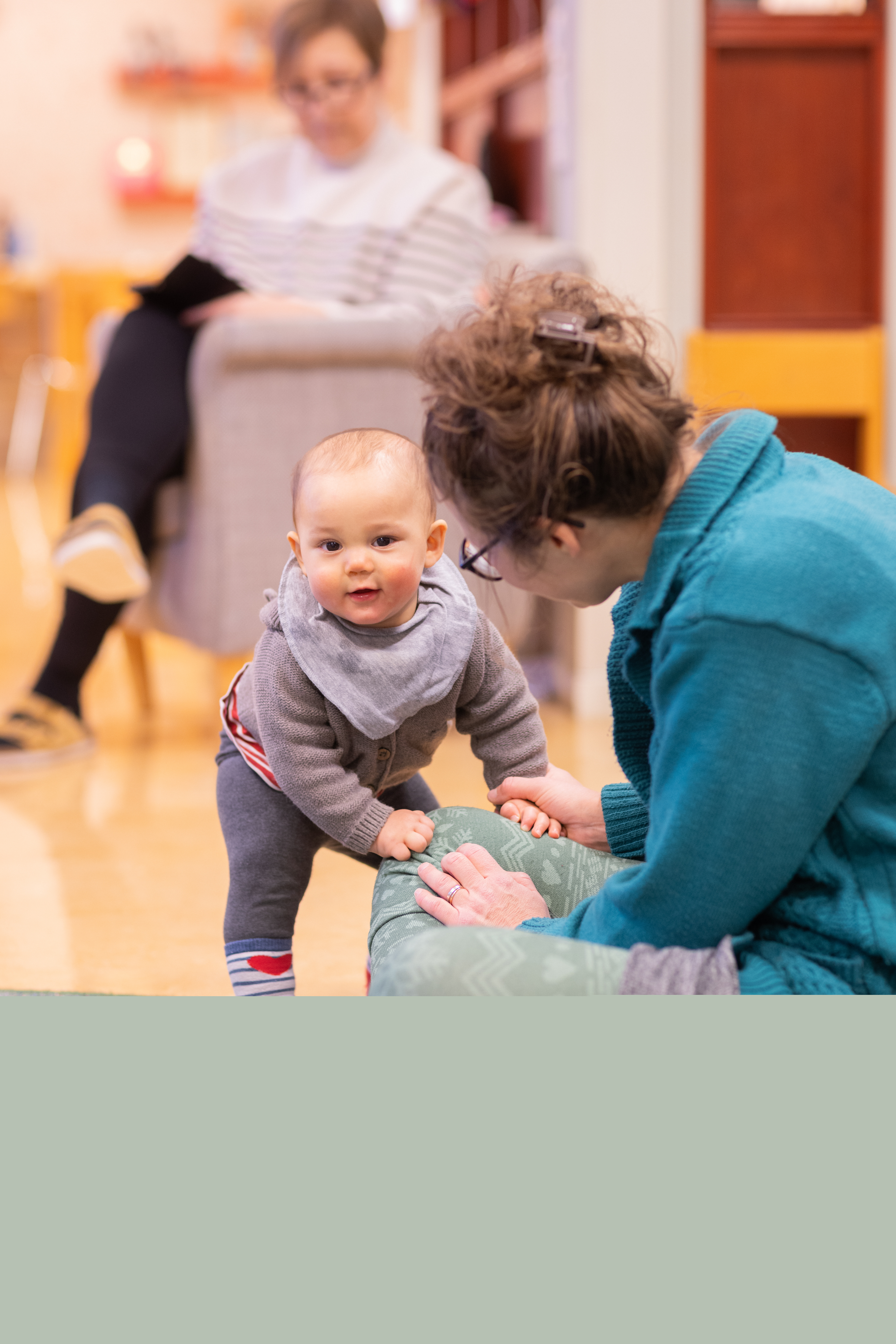 En bebis ställer sig trevande upp, stödd mot sin mamma.