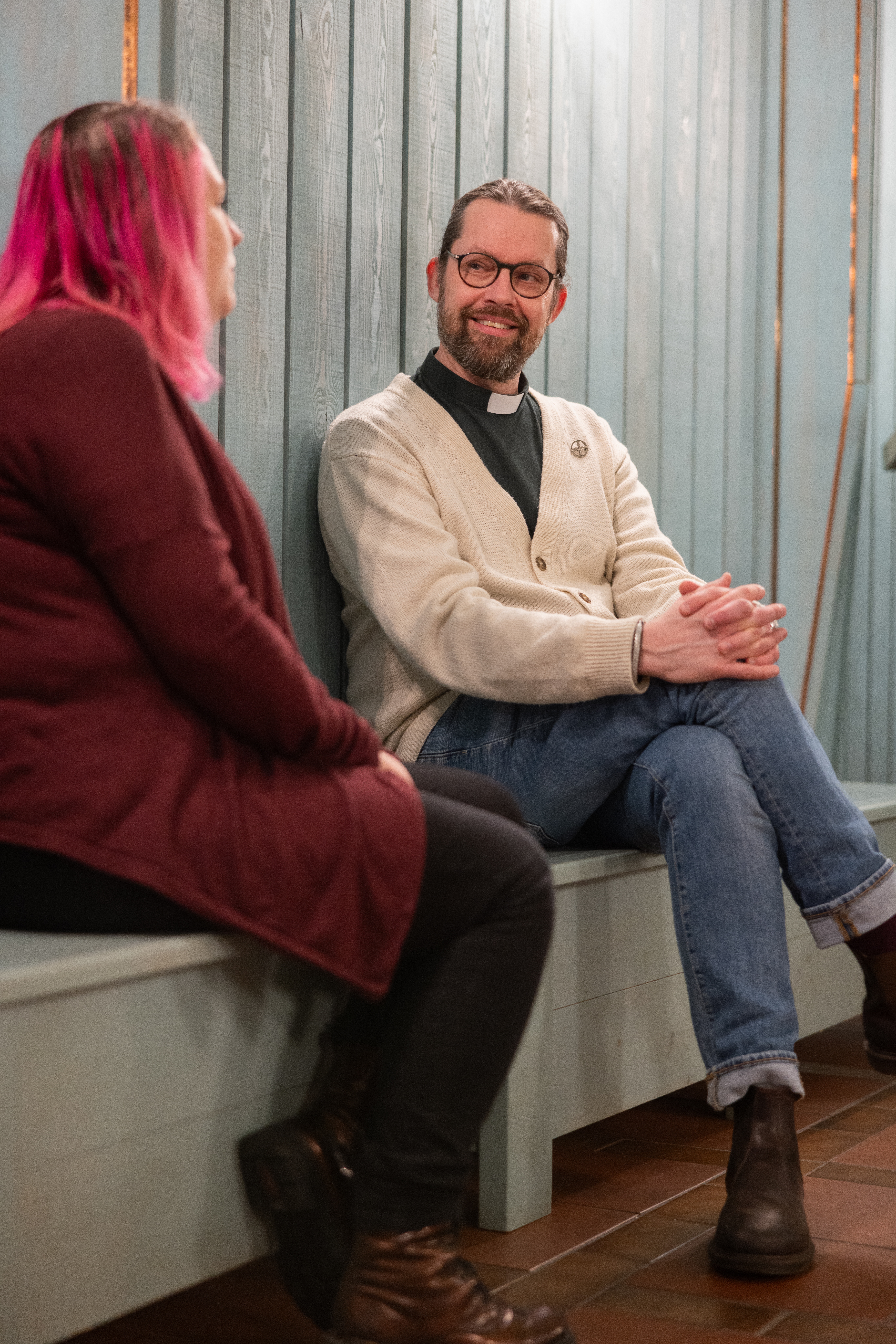 En diakon sitter på en träbänk inomhus och samtalar med en kvinna med rosafärgat hår.