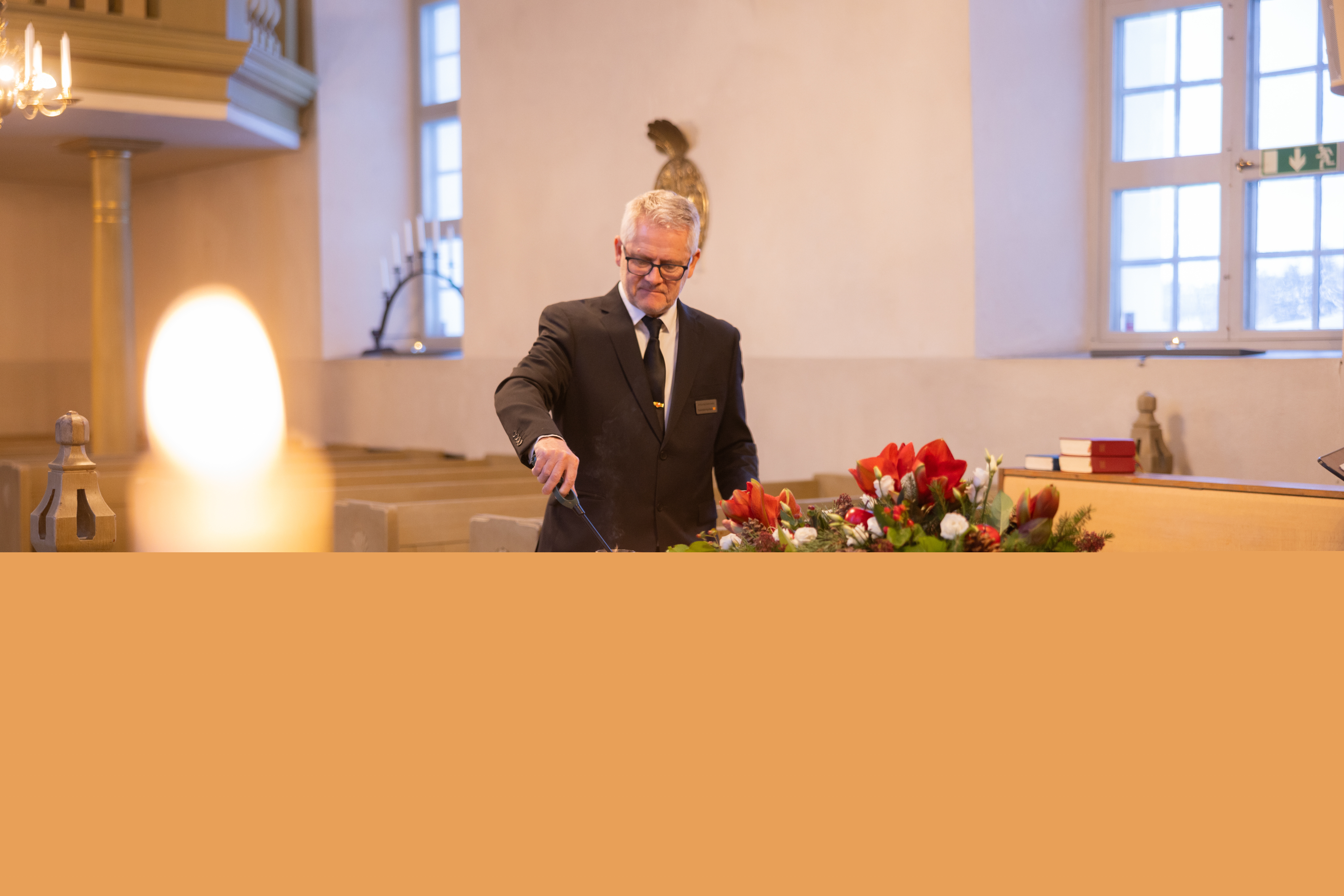 En kyrkvaktmästare tänder ett ljus på en vit kista som har en krans av amaryllis, tallkvistar och vita rosor.