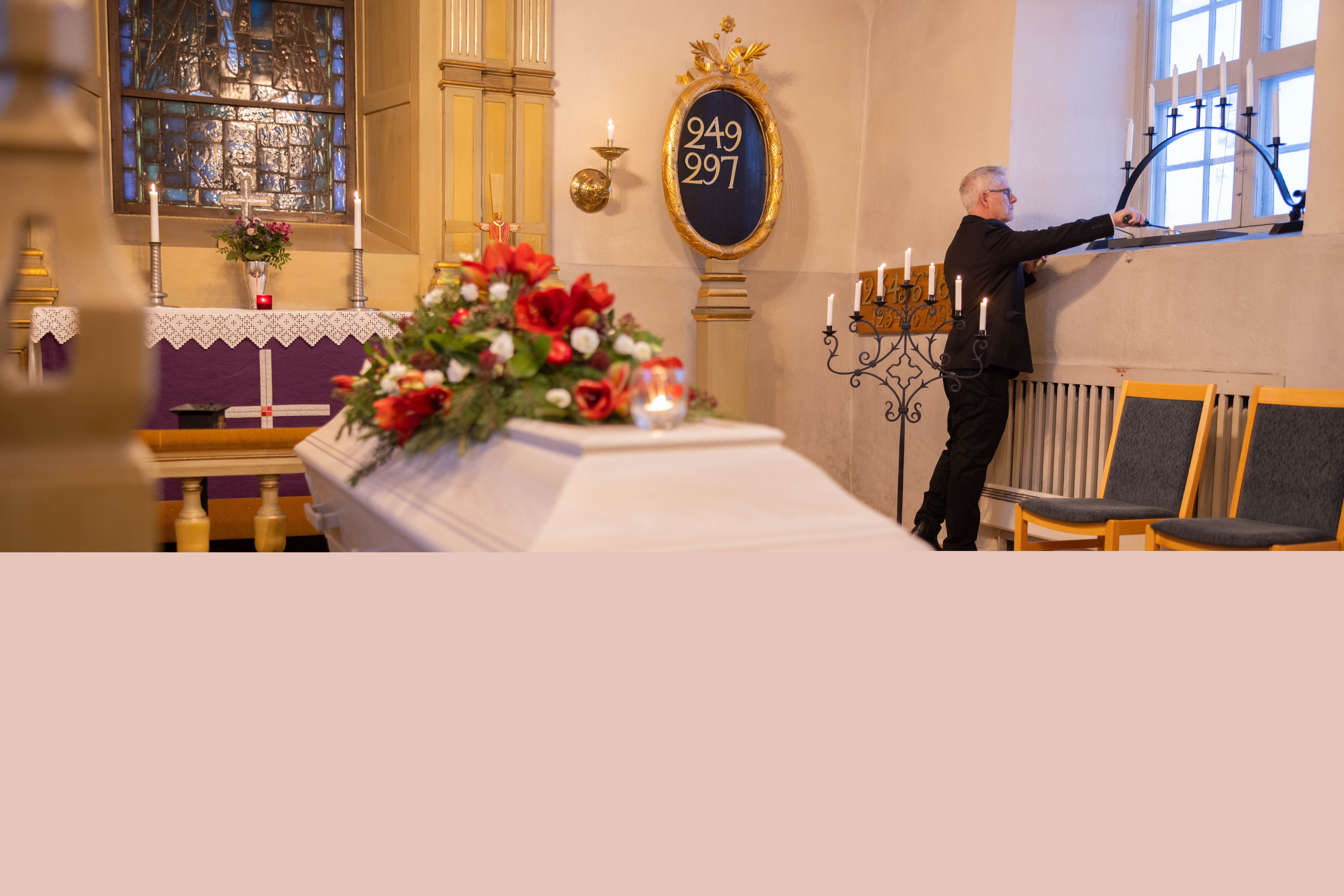 En kyrkvaktmästare tänder ljus i ett fönster. I koret står en vit kista med röda och vita blommor på.