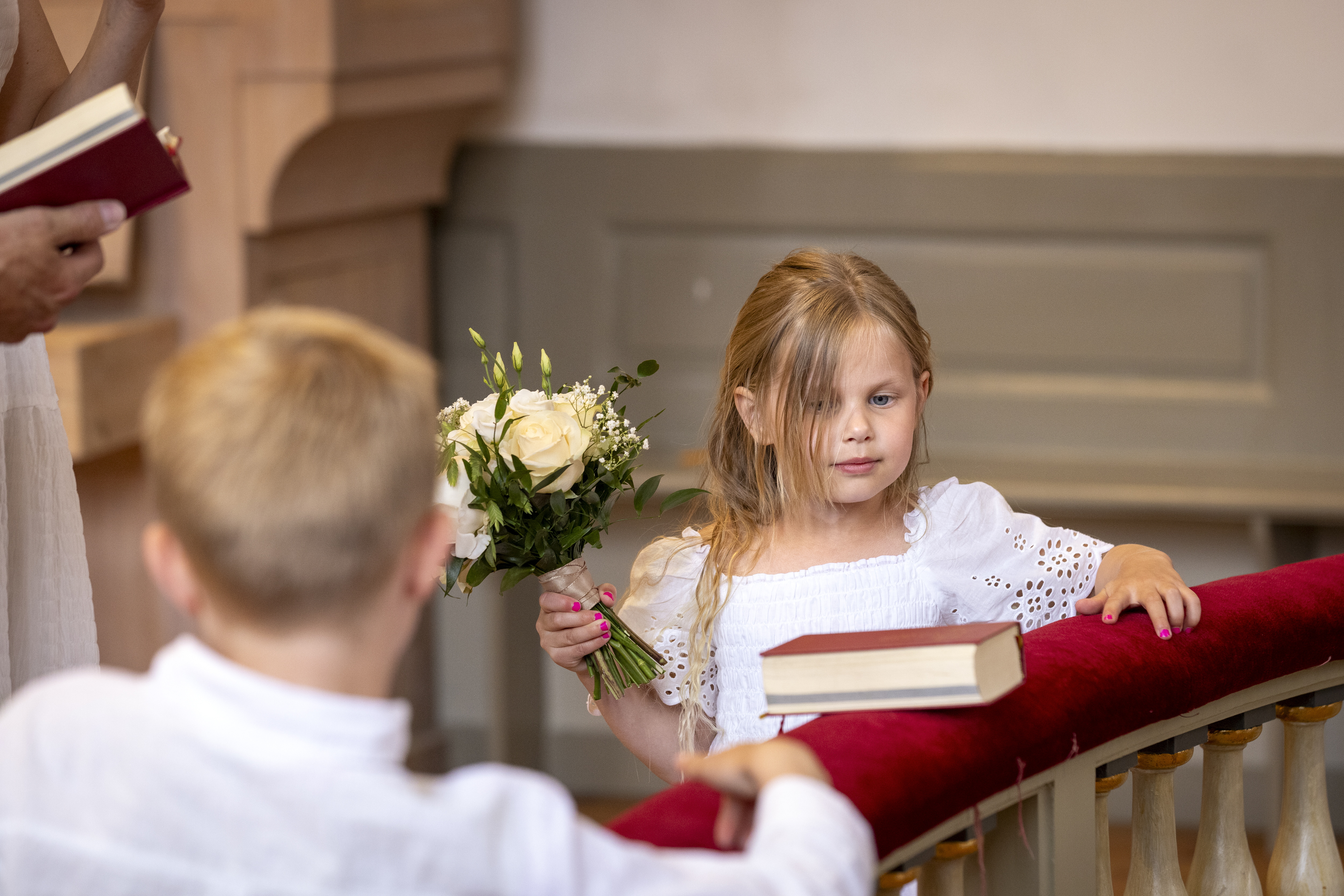 Två vitklädda barn står vid en altarring. Ett av barnen håller i en vit blombukett. En psalmbok ligger på altarringen.