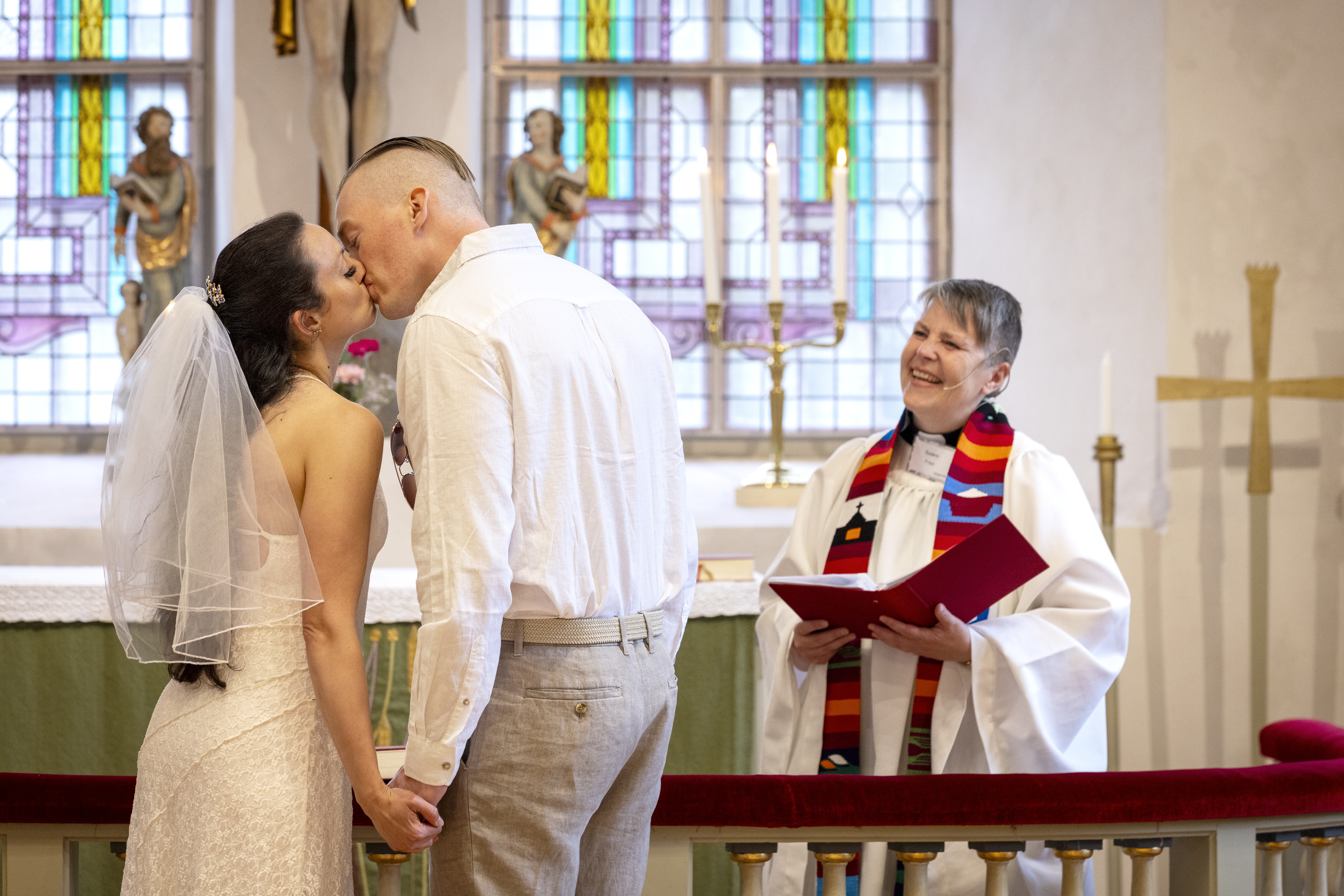 Ett brudpar kysser varandra framme vid altaret. En präst tittar på.