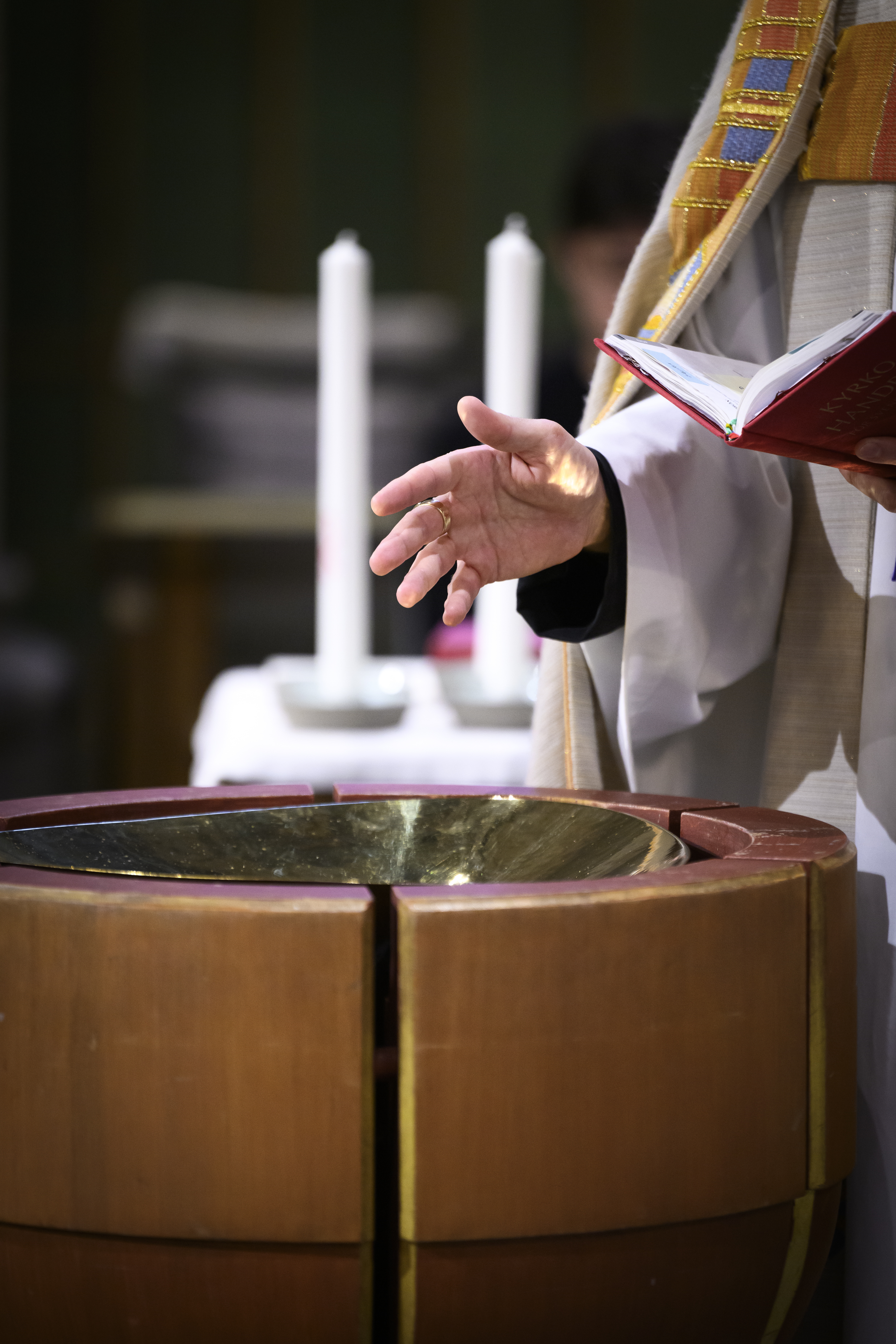 En präst står vid en dopfunt med en bibel i ena handen. Två dopljus står på ett bord i bakgrunden.