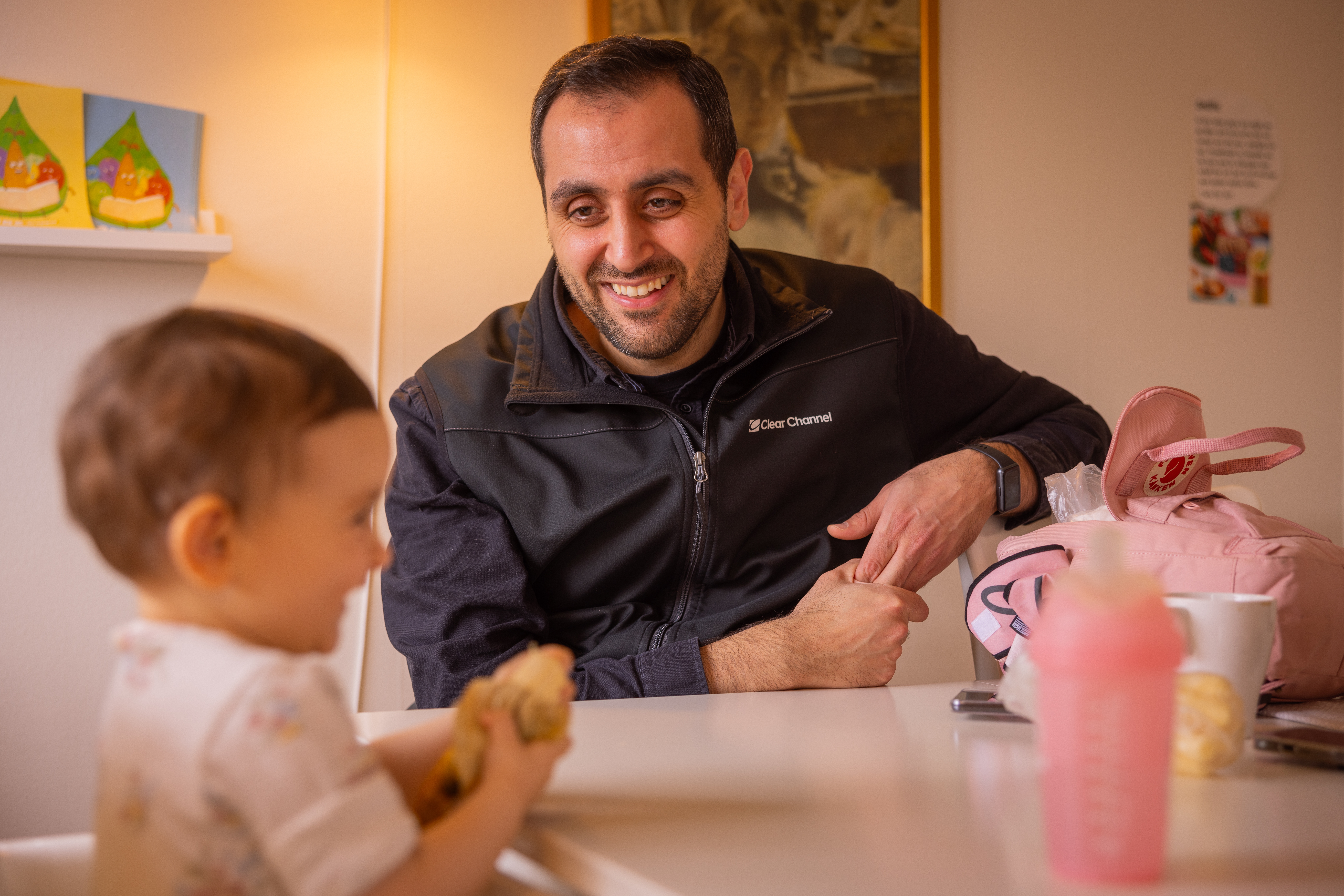 En leende man sitter vid ett bord och tittar på ett litet barn som sitter bredvid i en barnstol.