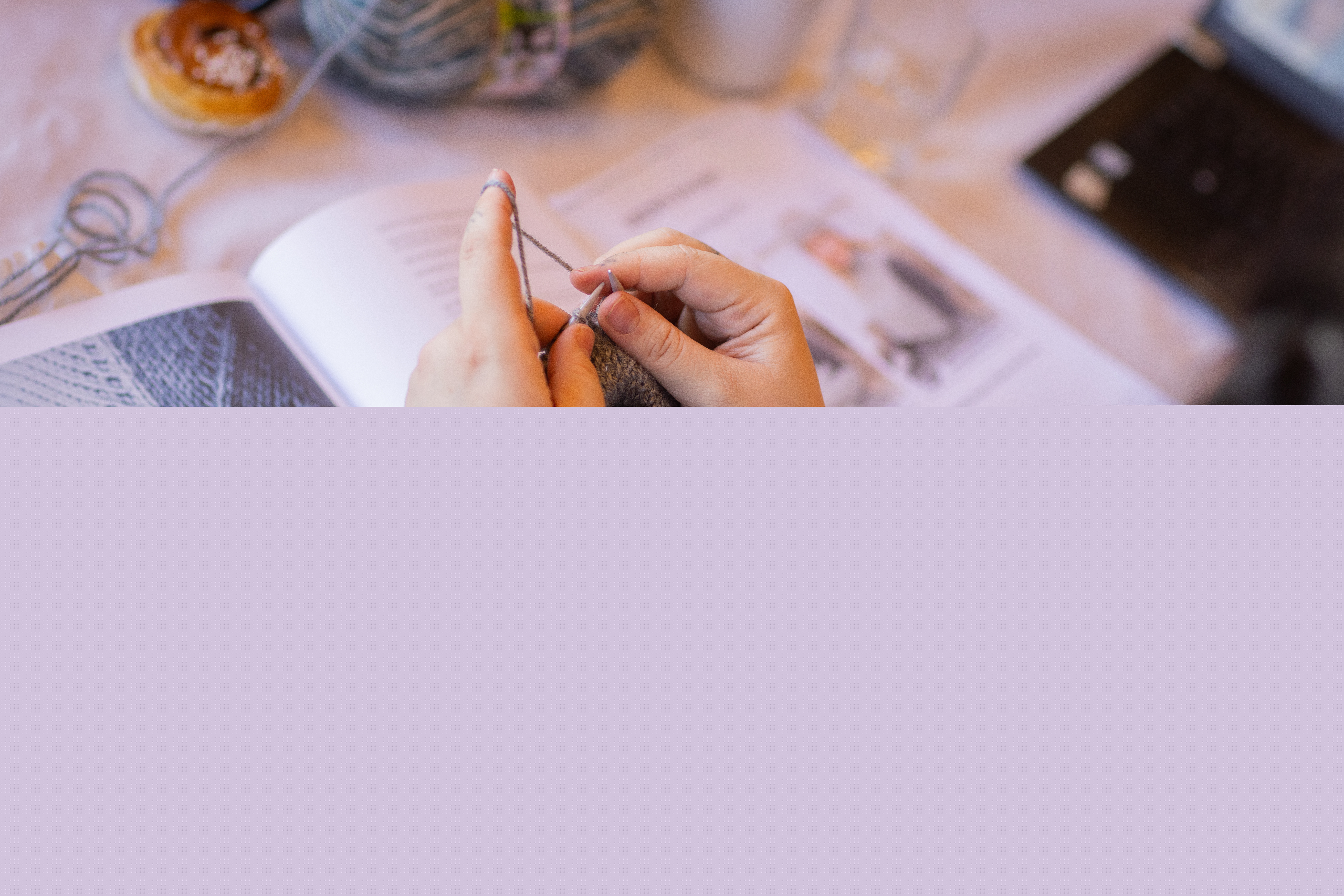 En person sitter vid ett bord och stickar. På bordet ligger en instruktionsbok i stickning.