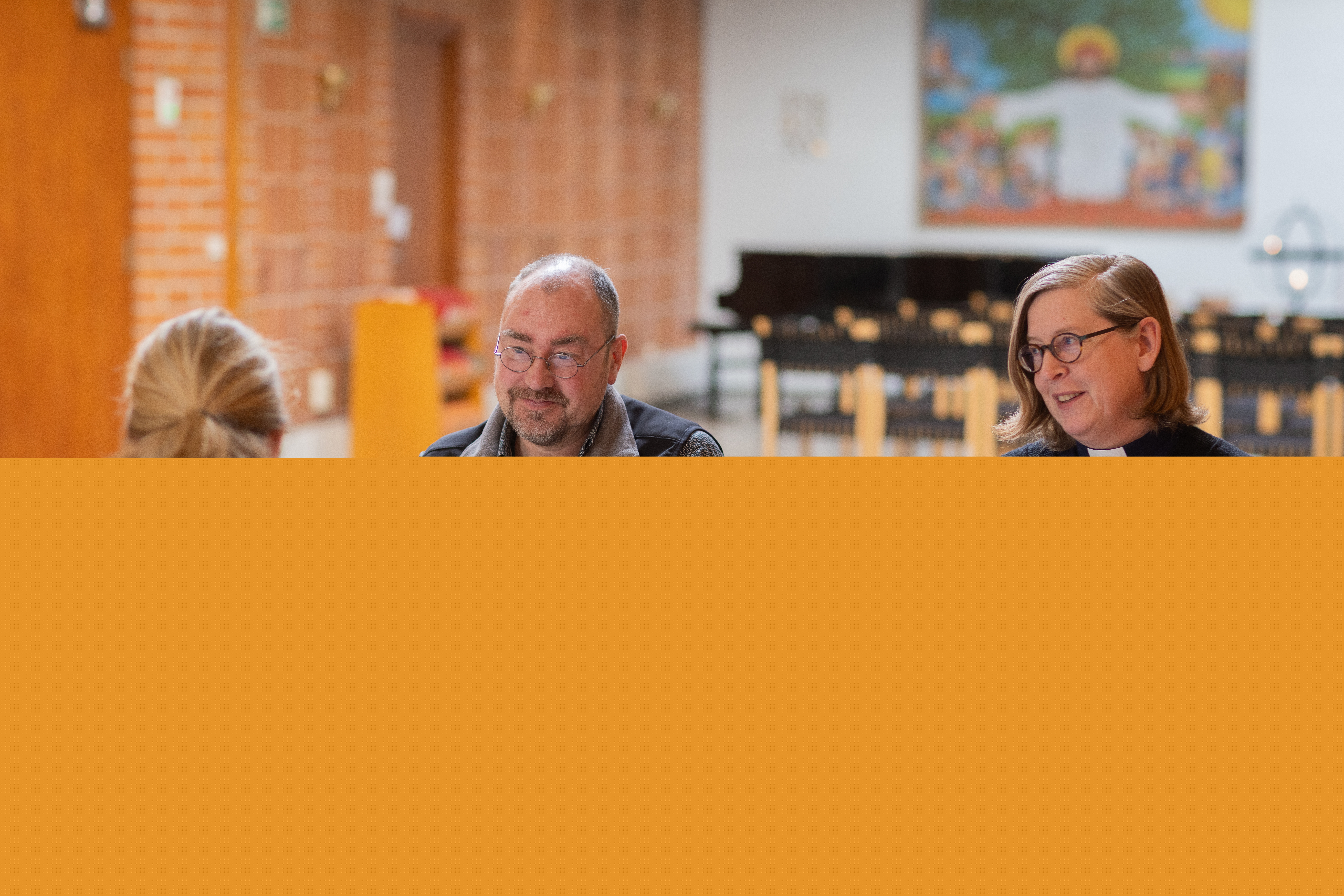 En präst sitter vid ett bord och samtalar med två andra personer.