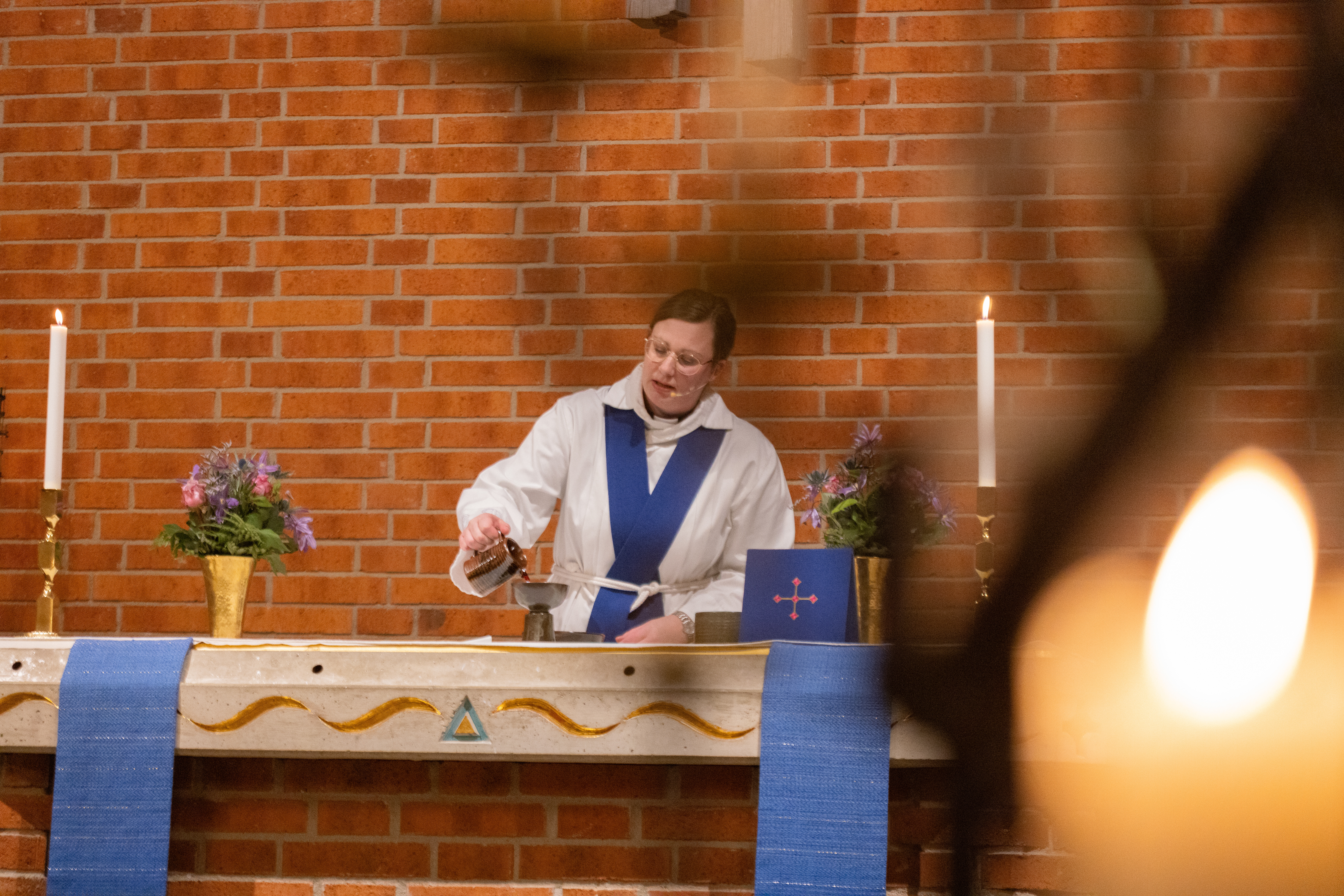 En kvinnlig präst med headset står vid ett altare och häller upp nattvardsvin. Tända ljus och blommor i guldfärgade vaser står på altaret.