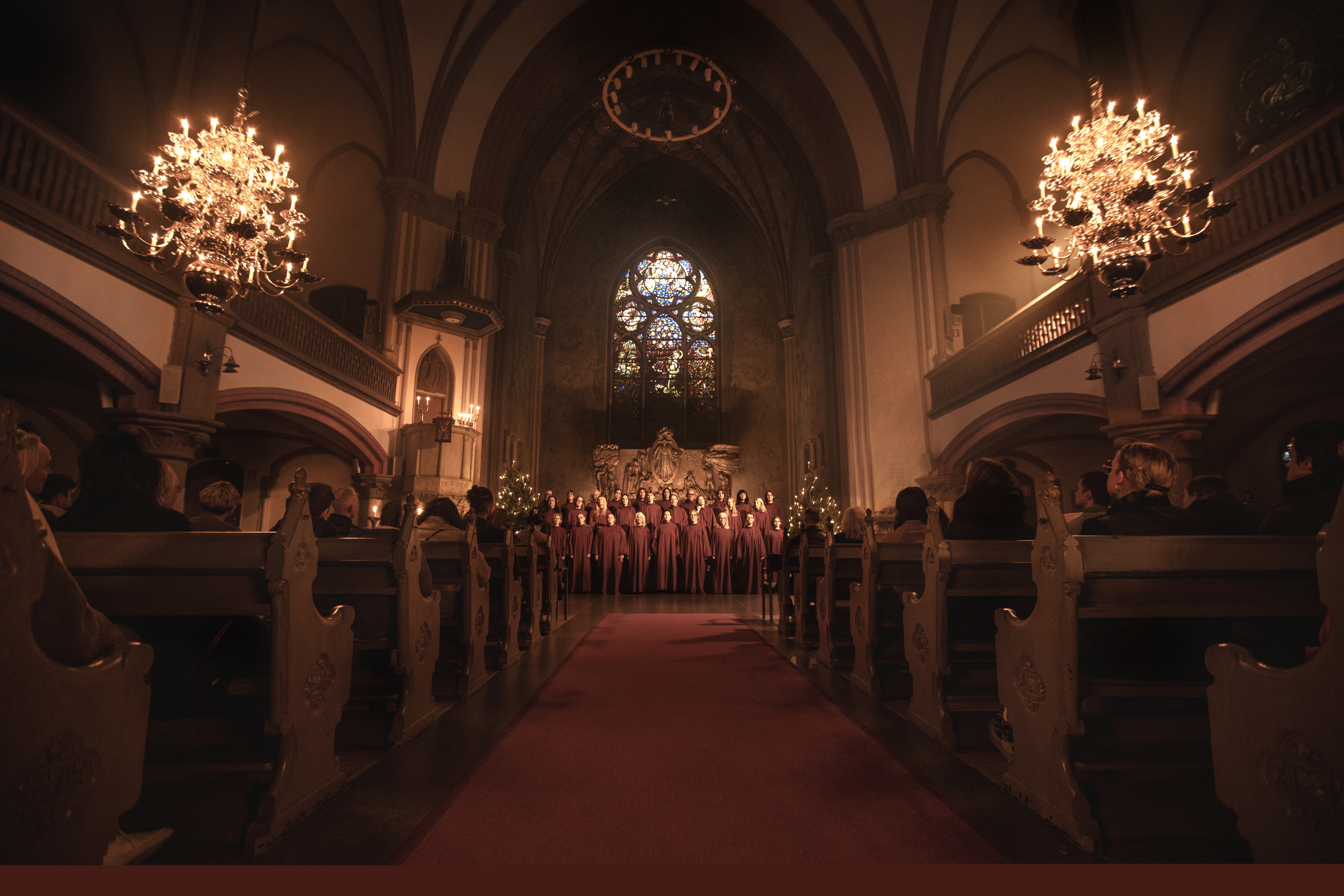 En altargång med kyrkbänkar och två upplysta ljuskronor på varsin sida. En kör står i koret.