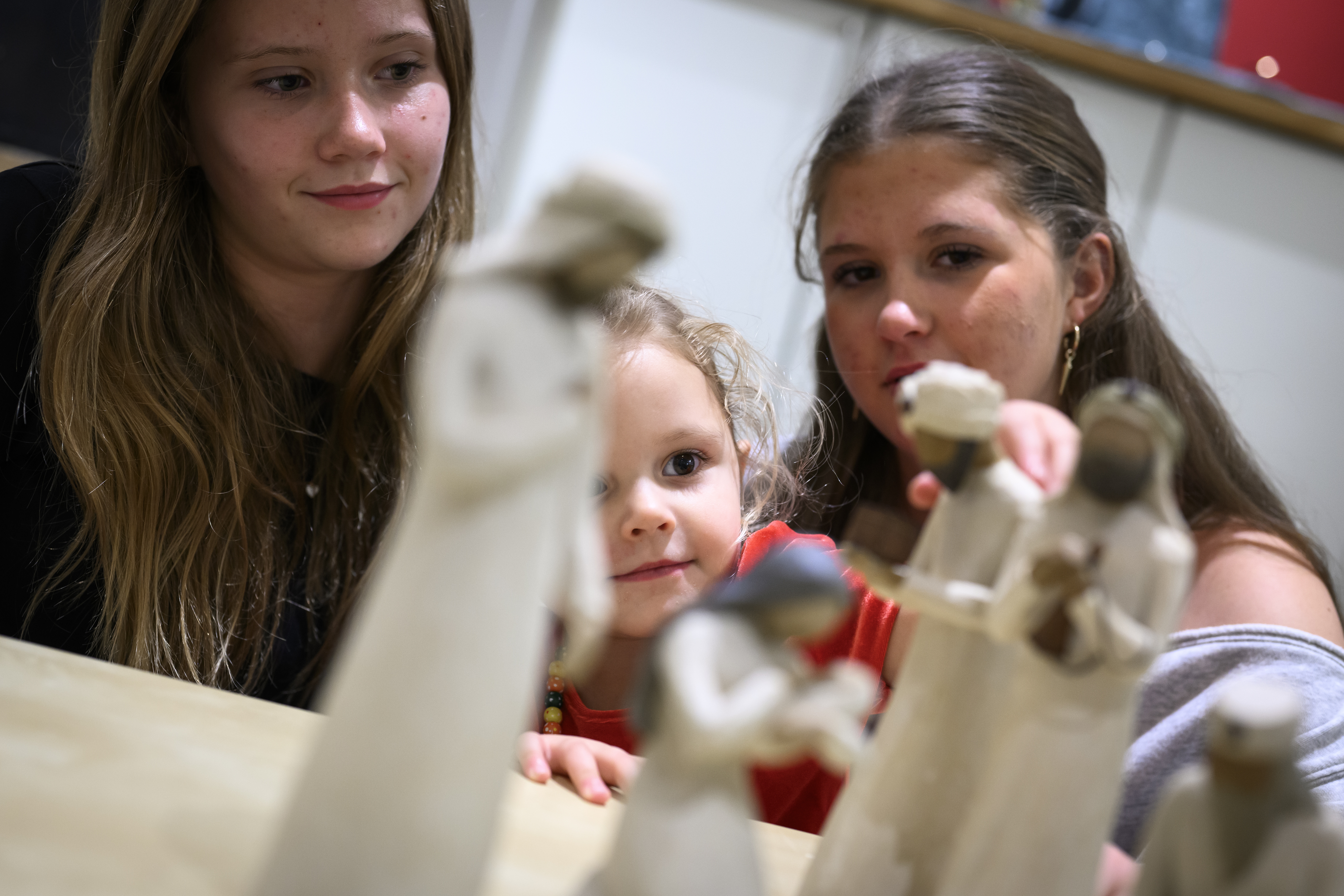 Trädockor som föreställer Jungfru Maria med Jesus-barnet i famnen och de tre vise männen står på ett bord. Ett barn och två ungdomar sitter runt bordet.