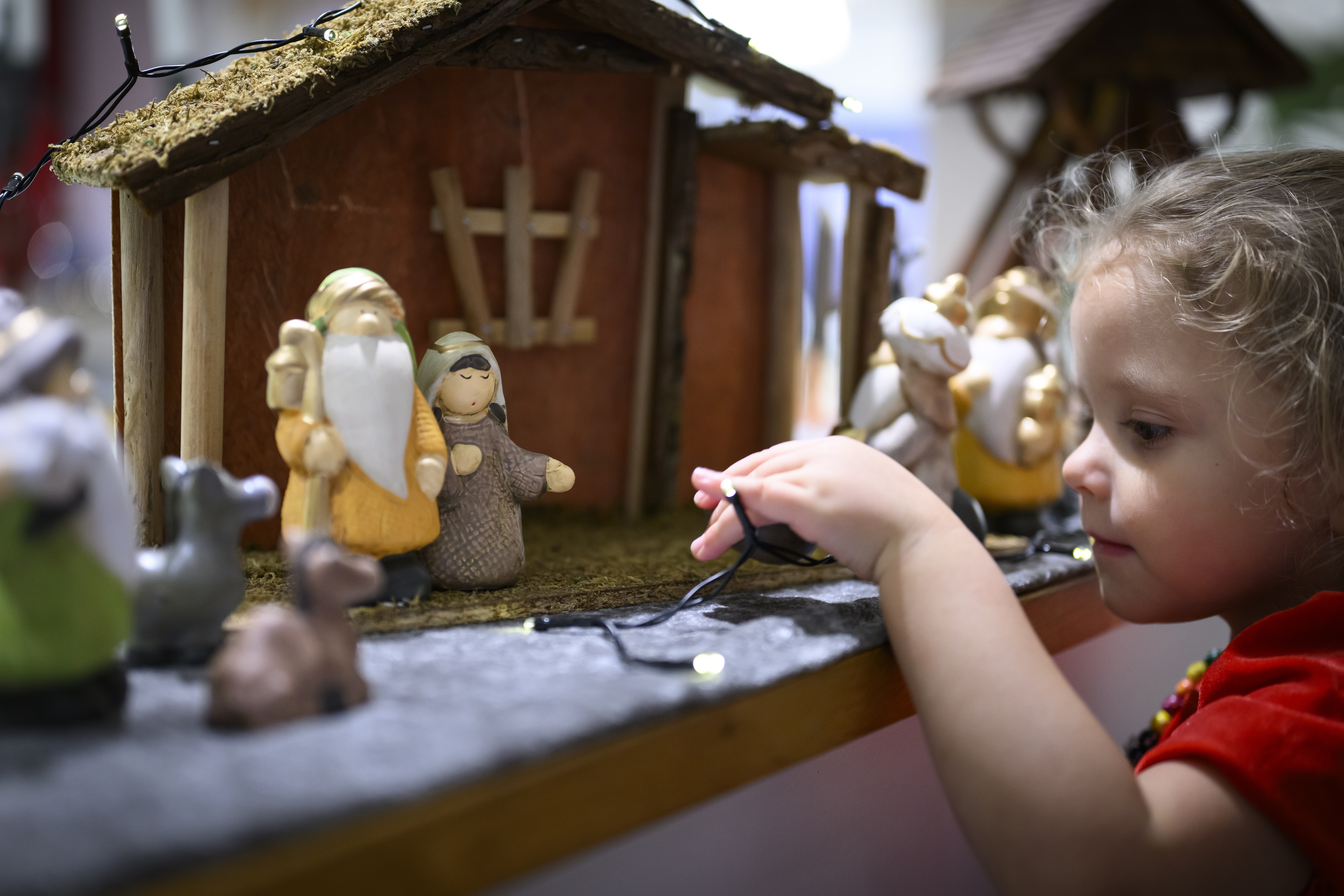 Ett litet barn håller en ljusslinga med ena handen. En julkrubba i bakgrunden.