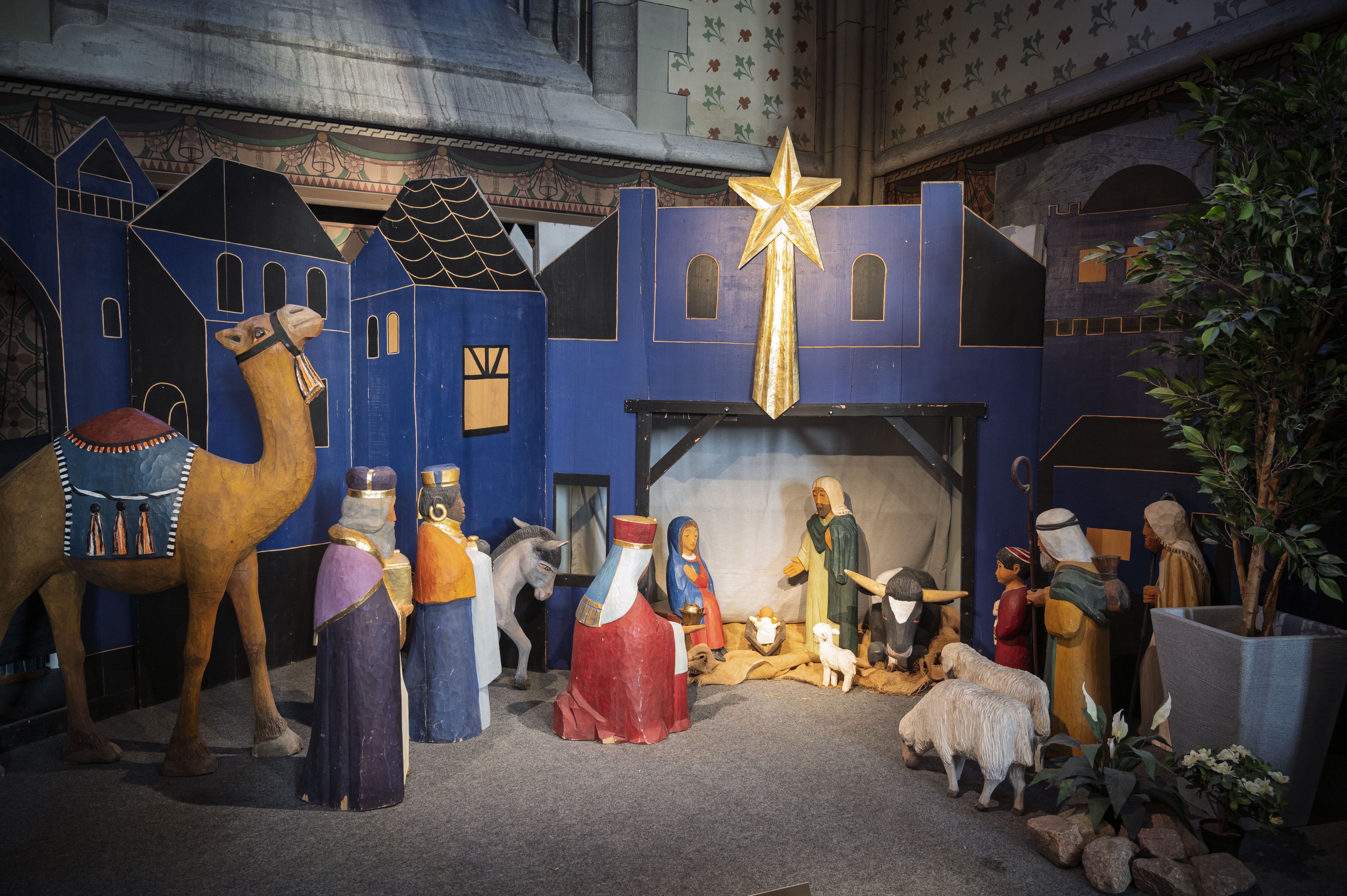 En julkrubba i ett blått hus med en stjärna på. Trädockor som föreställer de tre vise männen och en kamel i förgrunden.