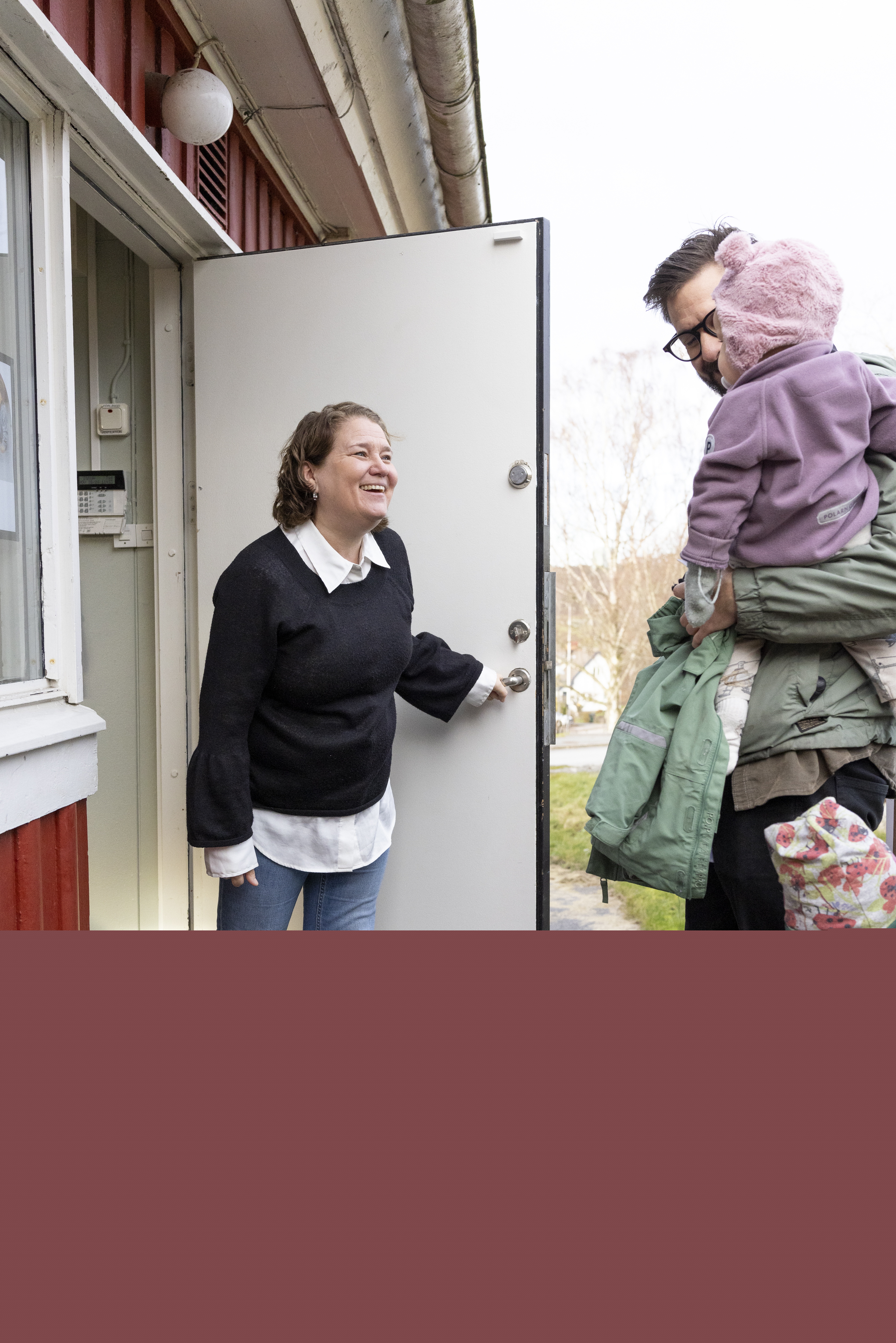 En kvinna öppnar en ytterdörr och hejar på en man och två barn.